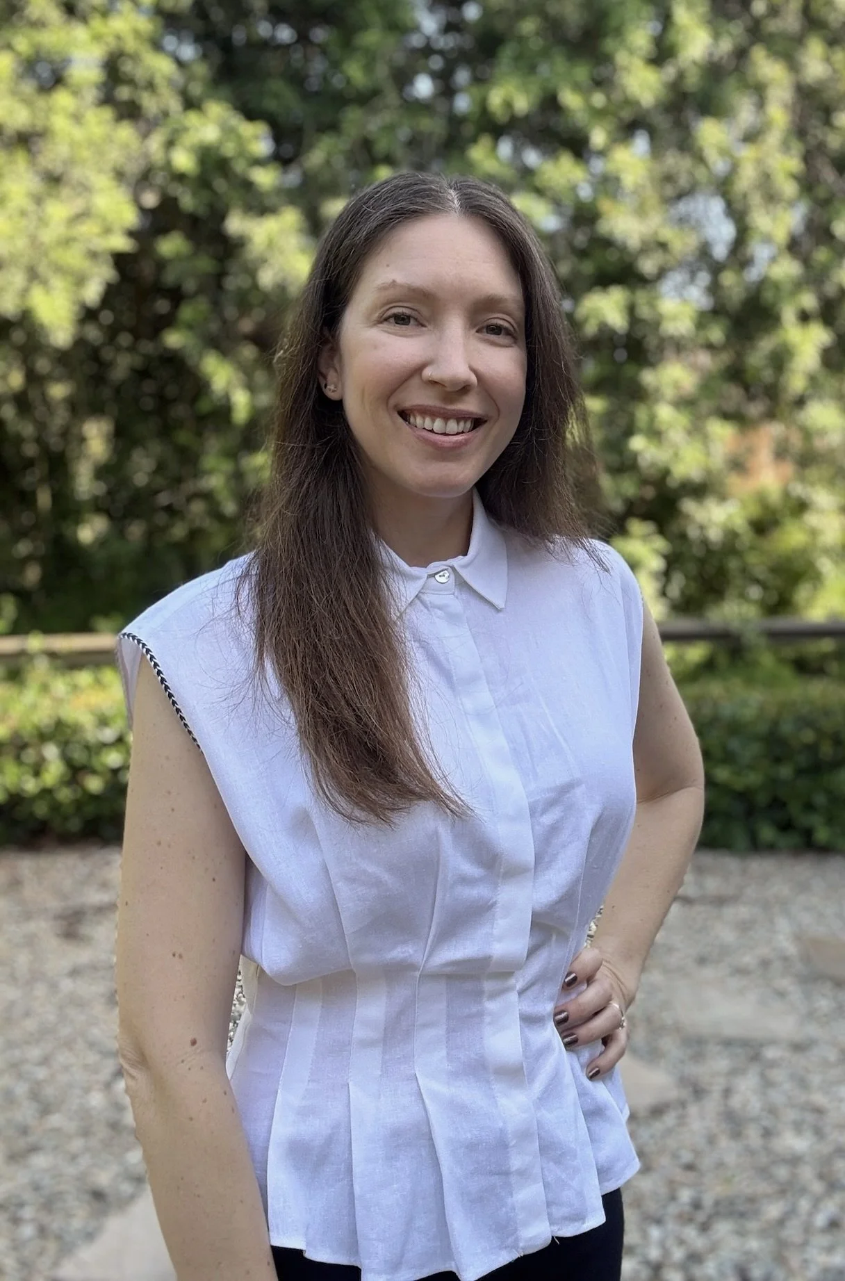 A woman with long brown hair, smiling, wearing a sleeveless white button-up top, standing outdoors with trees and greenery in the background.