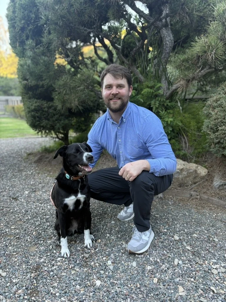 A man with a beard wearing a blue button-up shirt and black pants kneeling next to a black and white dog on a gravel path in a garden with green trees and plants.