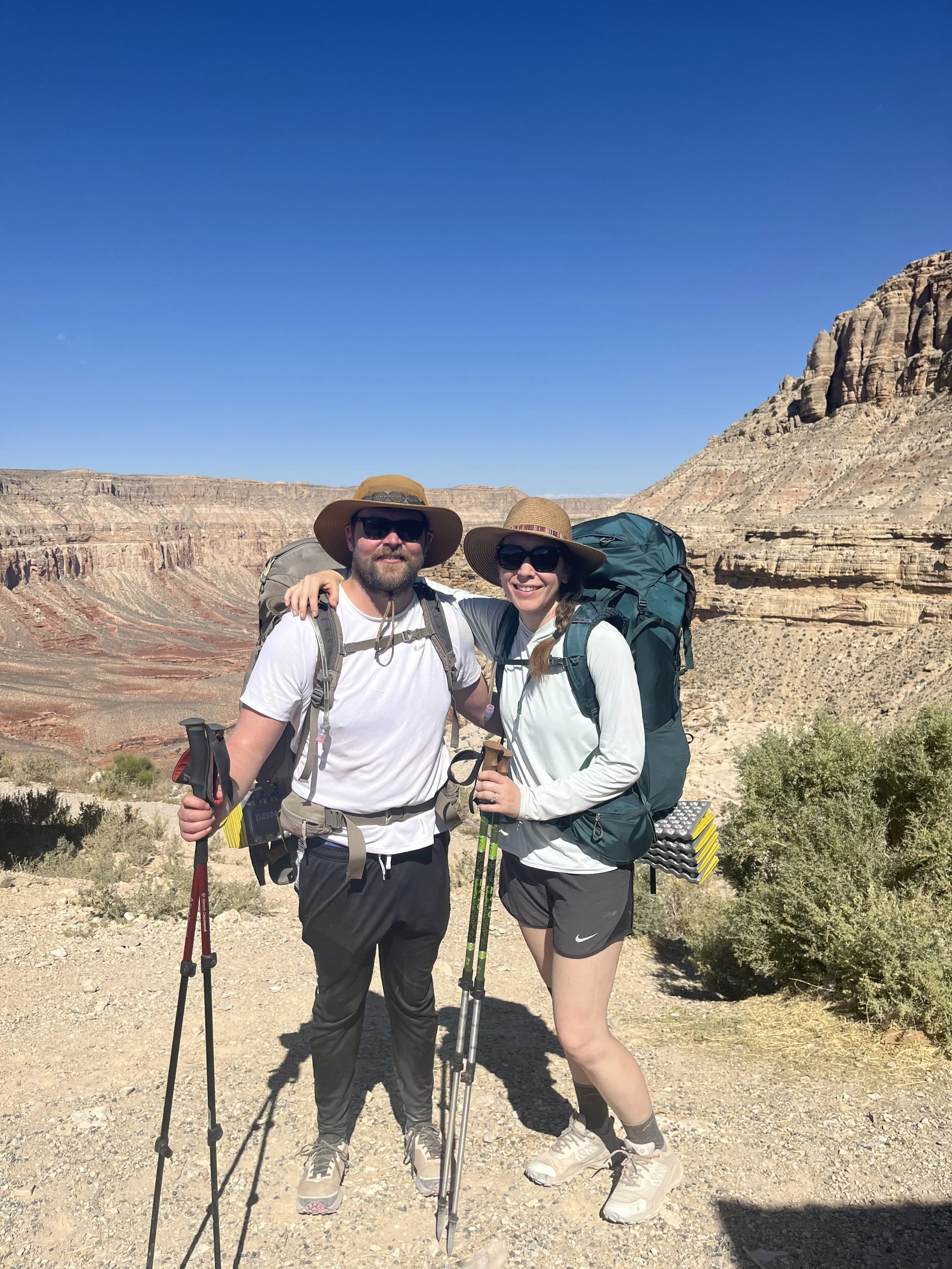 Two hikers, a man and a woman, standing together outdoors in a canyon with layered rock formations in the background. They are smiling, wearing hats, sunglasses, and backpacks, holding trekking poles, dressed in outdoor clothing suitable for hiking.