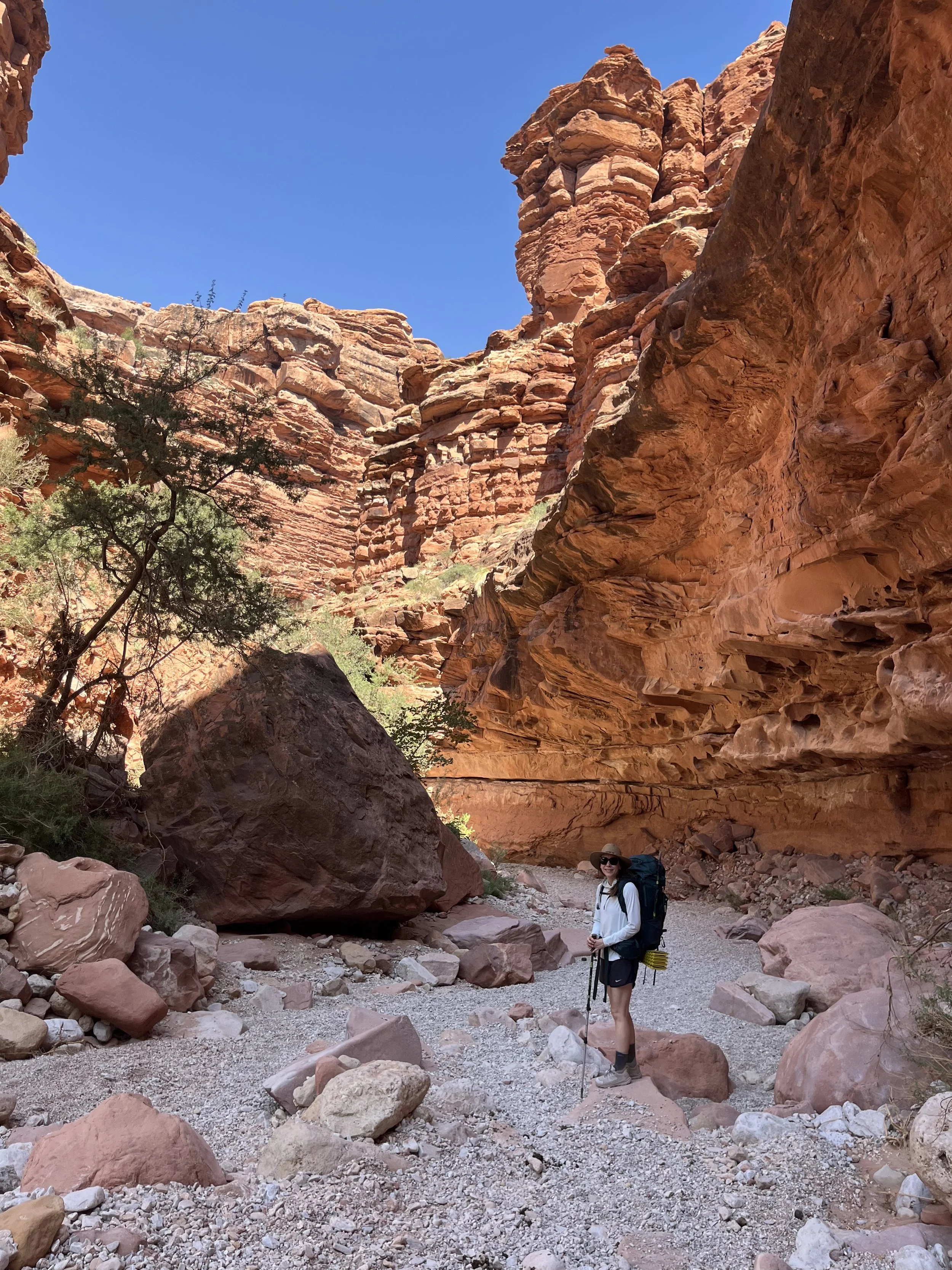A woman standing with a backpack and hiking pole in a rocky canyon with red sandstone cliffs and a clear blue sky.