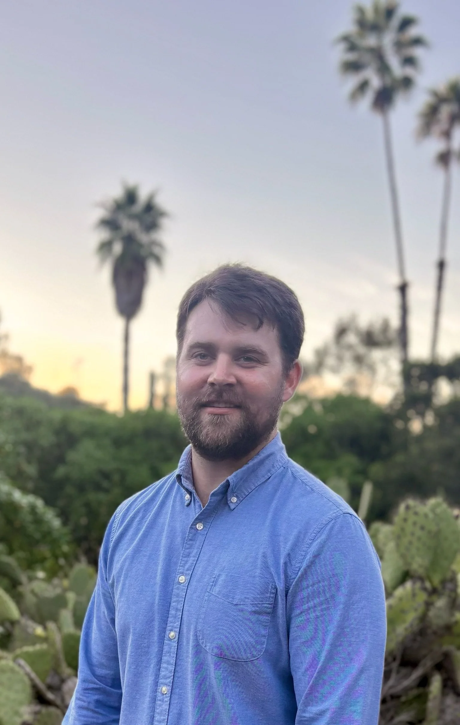 A man wearing a blue button-up shirt standing outdoors with palm trees and greenery in the background at sunset.