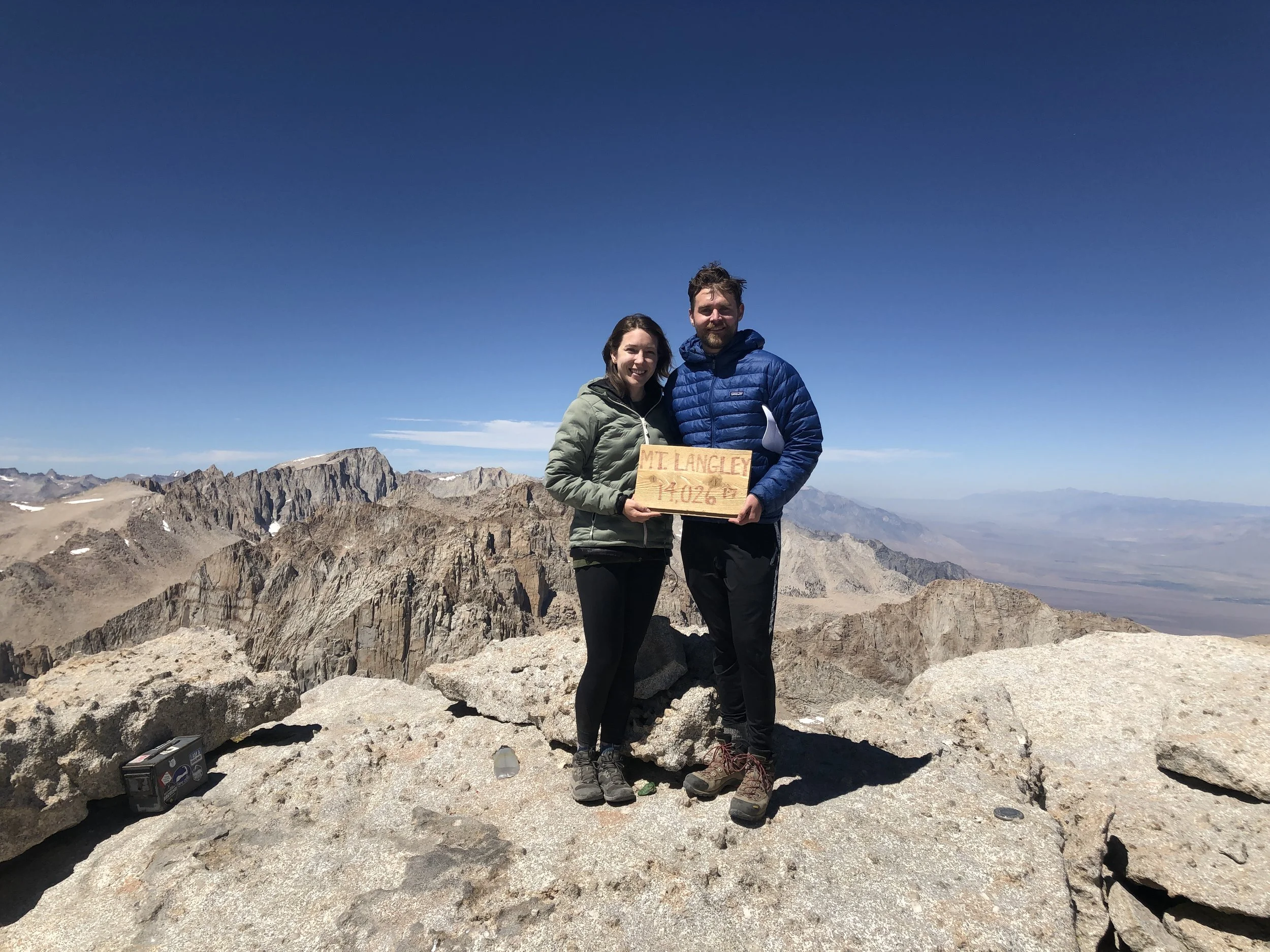 Two hikers at the summit of Mount Langley, standing on rocky terrain with a mountain range and blue sky in the background. They are smiling and holding a wooden sign that reads "Mt. Langley 14,042 ft".