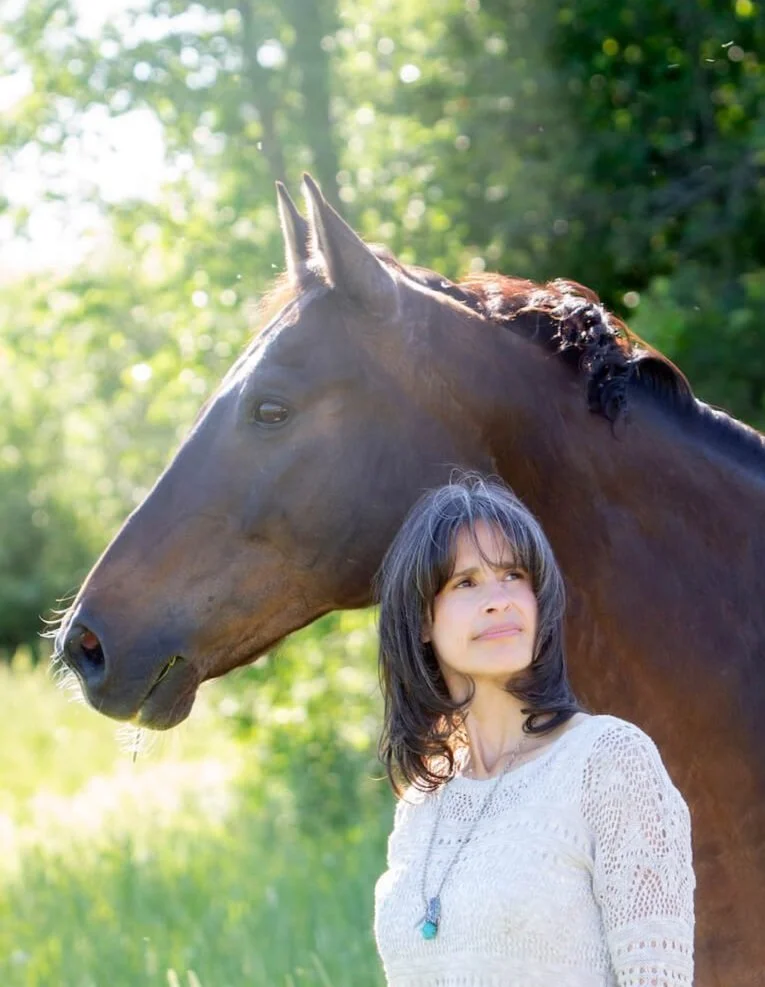 Une femme avec des cheveux noirs et une robe blanche, devant un cheval brun dans un paysage verdoyant au soleil.