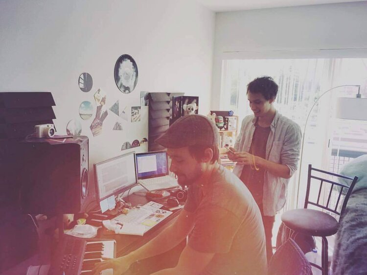 Two people in a home studio setup, one playing a keyboard, surrounded by monitors, speakers, and wall decorations. Sunlight enters through a window, illuminating the room.