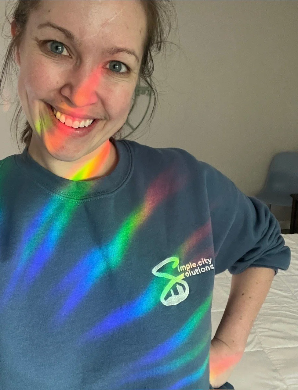 A smiling sarah stands in a blue-Simple.city Solutions-logoed sweatshirt. A prism reflects rainbows on her.
