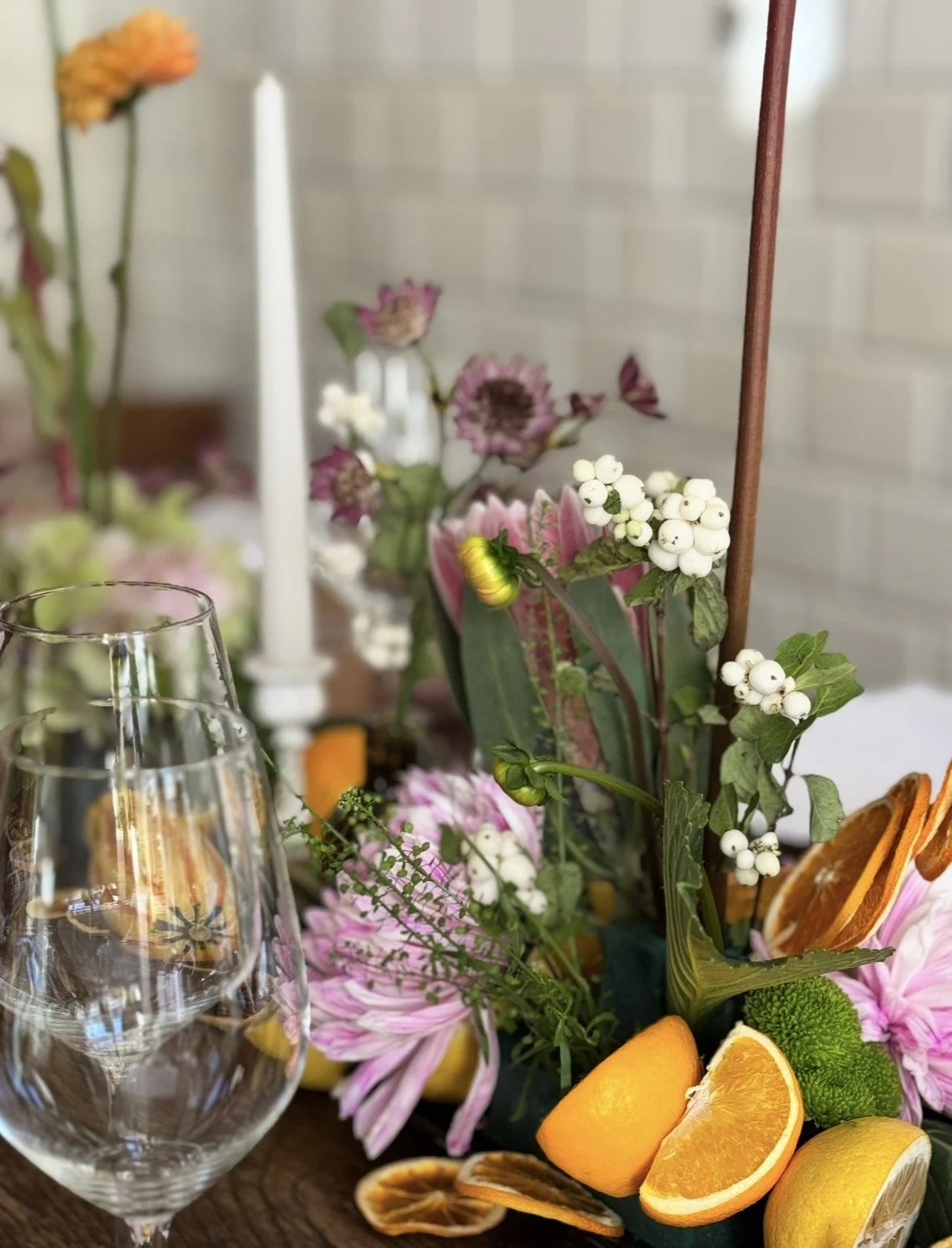 Blumenstrauß mit bunter Blumendekoration, Orangen und Zitronen auf einem Tisch, Weingläser im Vordergrund, Kerzen im Hintergrund.