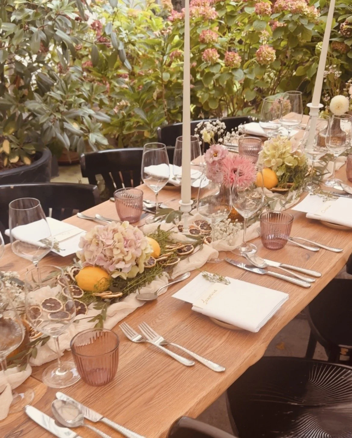 Ein schön eingedeckter Tisch mit Kerzen, Blumen und Gläsern, im Hintergrund eine Gartenumgebung mit Pflanzen und blühenden Sträuchern.