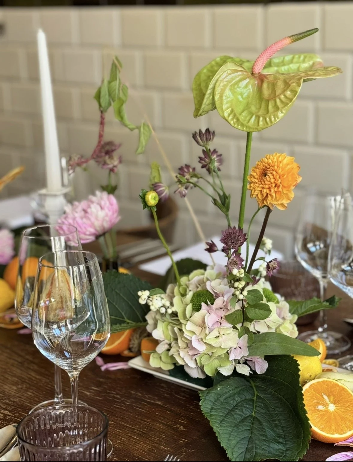 Blumenarrangement auf einem Holztisch, mit Hydrangea, Chrysanten, weiteren Blumen und großen grünen Blättern, umgeben von Gläsern, Orangen und Kerzen.