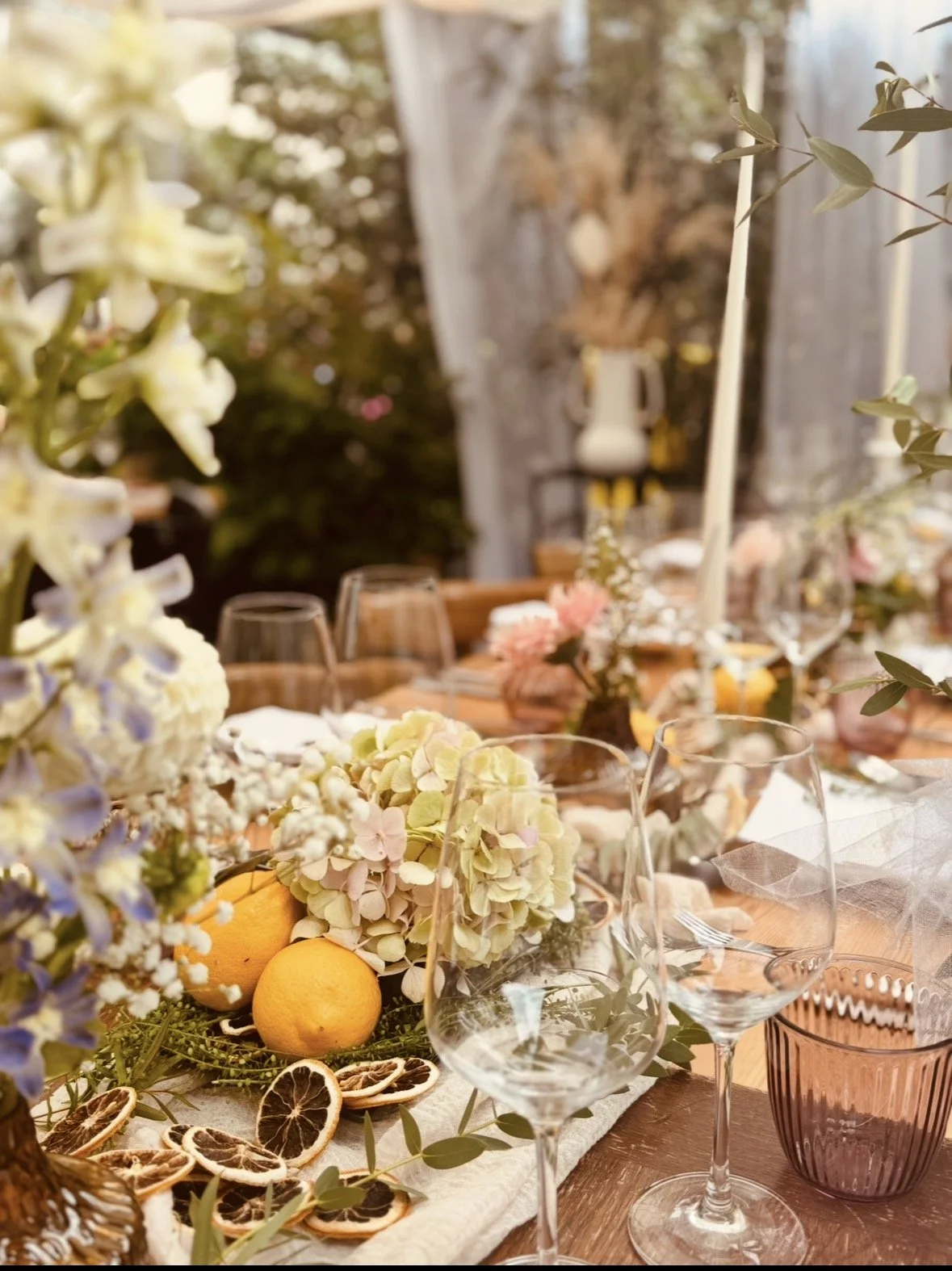 Ein festlich gedeckter Tisch mit Blumen, Zitronen, getrockneten Zitrusfrüchten und leeren Weingläser, im Hintergrund eine gemütliche Outdoor-Umgebung mit Pflanzen und Dekorationen.