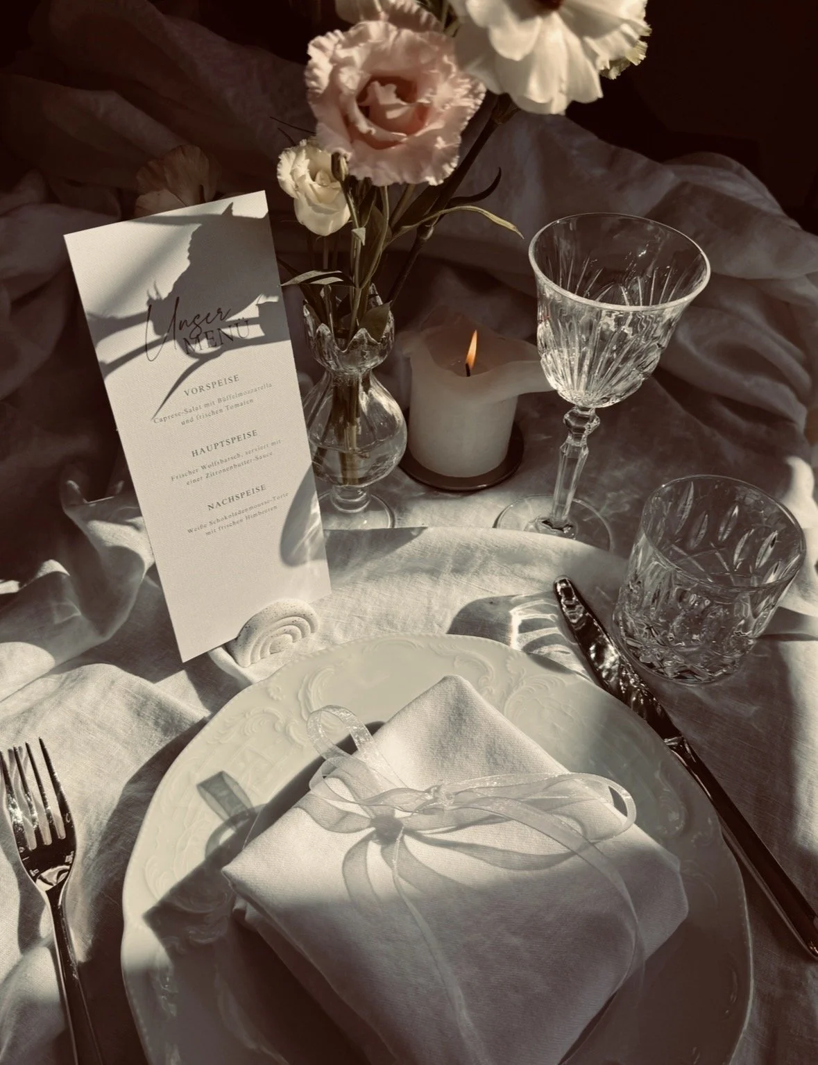 Ein eleganter gedeckter Tisch mit weißen Tellern, Besteck, Kristallgläsern, einer Kerze, einem Blumenstrauß und einer Menükarte. Die Atmosphäre wirkt stilvoll und festlich.