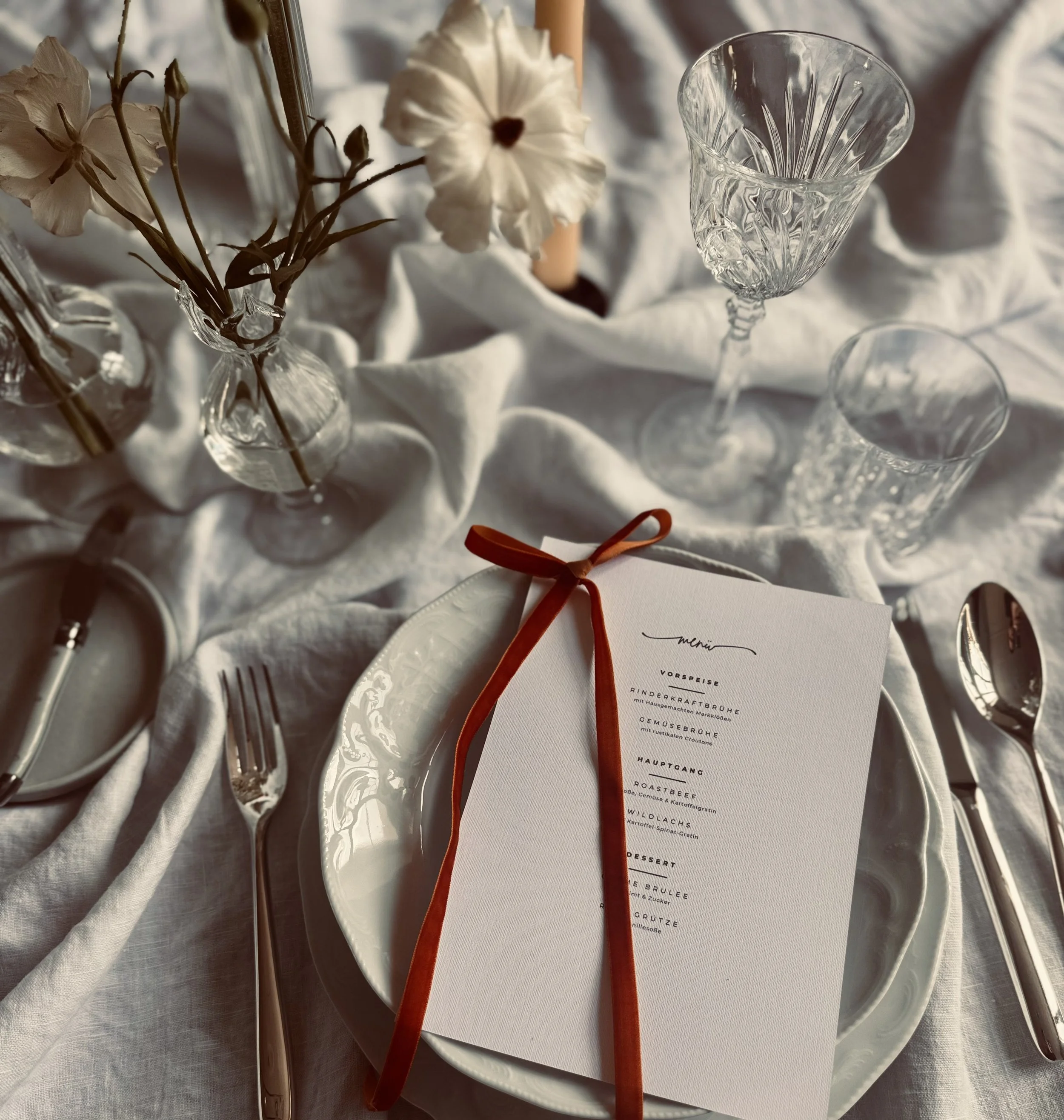 Eine festliche Tafel mit einem weißen Leinentisch, Wein- und Wassergläser, Blumen in Kristallvasen, Menükarte mit roten Band, Besteck und eine gedeckte Tortenplatte im Hintergrund.