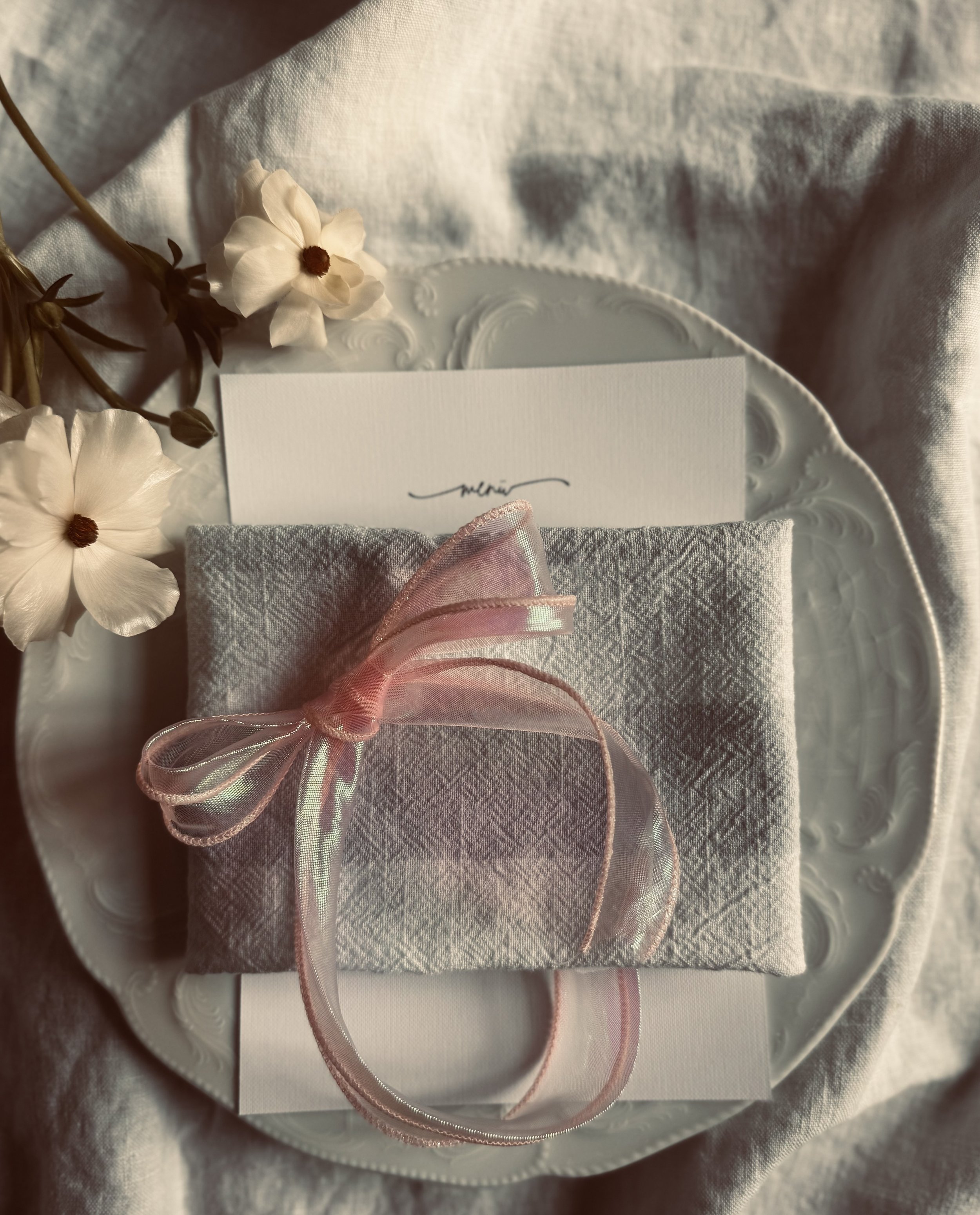 Ein Teller mit einer grau marmorierten Geschenkverpackung, verziert mit rosa transparentem Schleifenband, und weißen Blumen auf einem weißen Tuch.