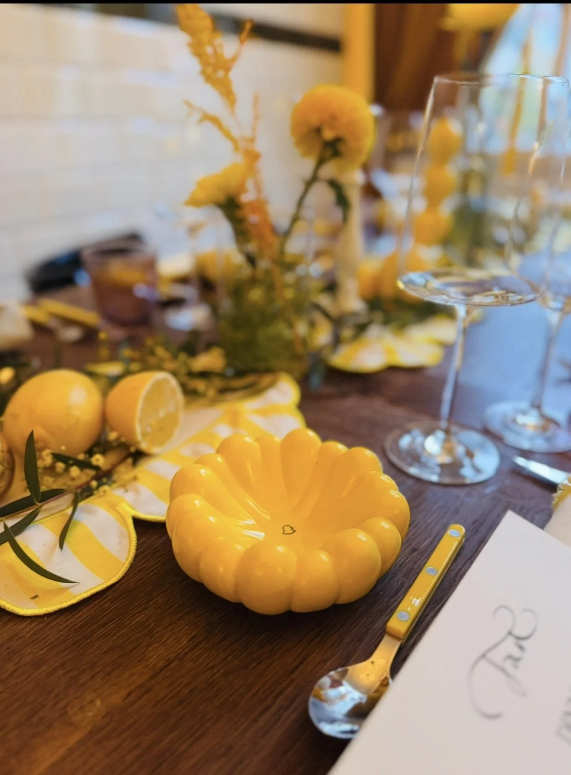Ein Tisch mit gelben Dekorationen, einschließlich einer gelben, mandelartigen Schale, Zitronen, gelben Blumen und gelben Besteckteilen, bei einer festlichen Gelegenheit.