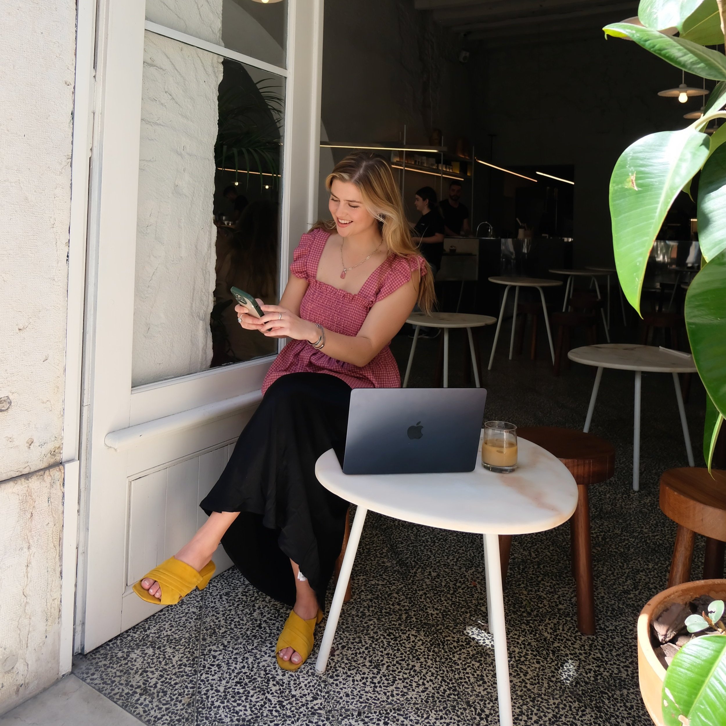 A young woman with long blonde hair is sitting on a chair outside a café, looking at her phone and smiling. She has a laptop and a glass of iced coffee on a small round table in front of her.