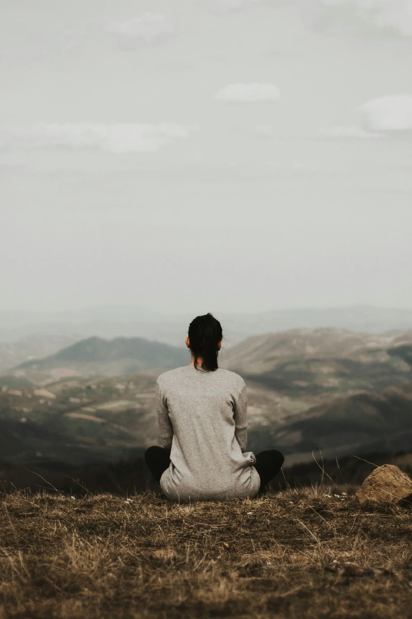 A person sitting cross-legged on a grassy hilltop, facing away, overlooking a vast landscape with mountains and cloudy sky.