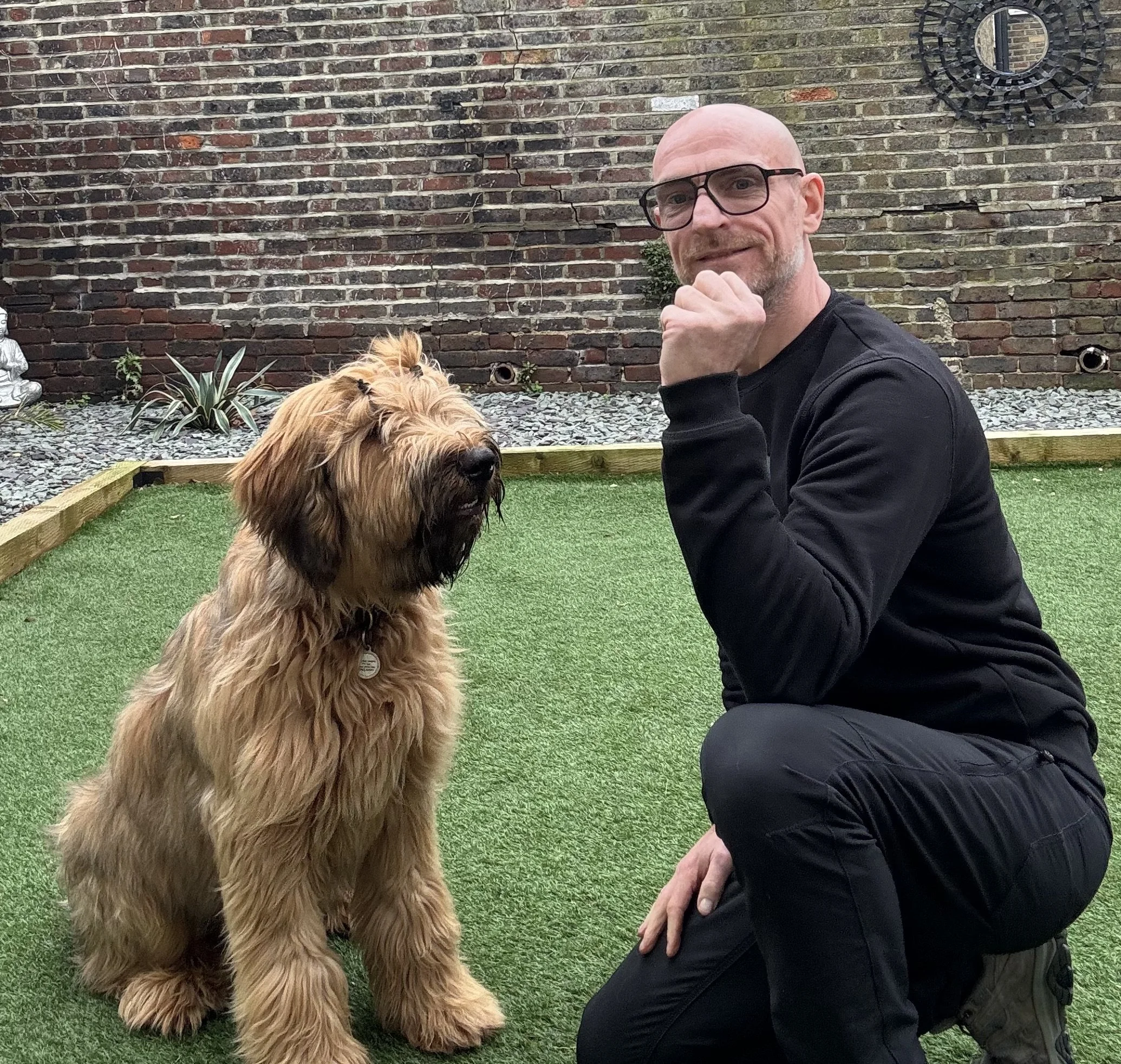 A man with glasses and a beard kneeling on one knee beside a large, fluffy, tan-colored dog in a backyard with a brick wall and small garden decorations.