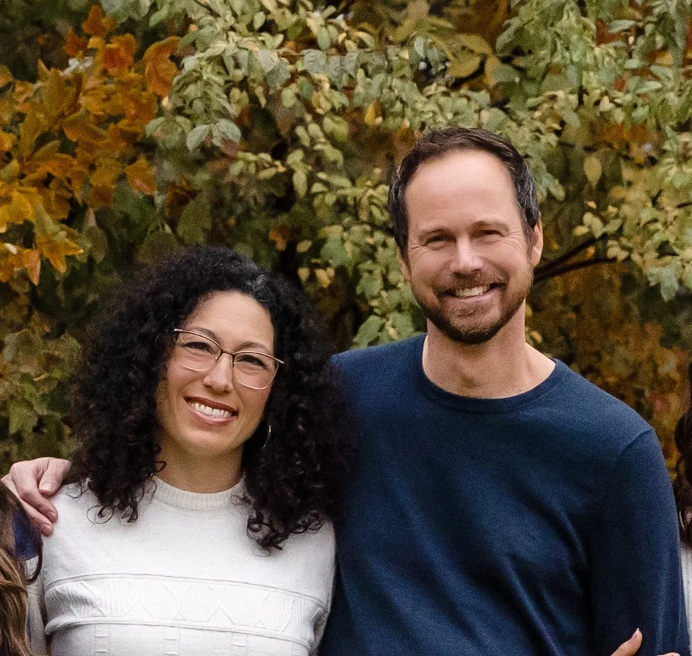 A man and woman standing outdoors among autumn foliage, smiling at the camera.