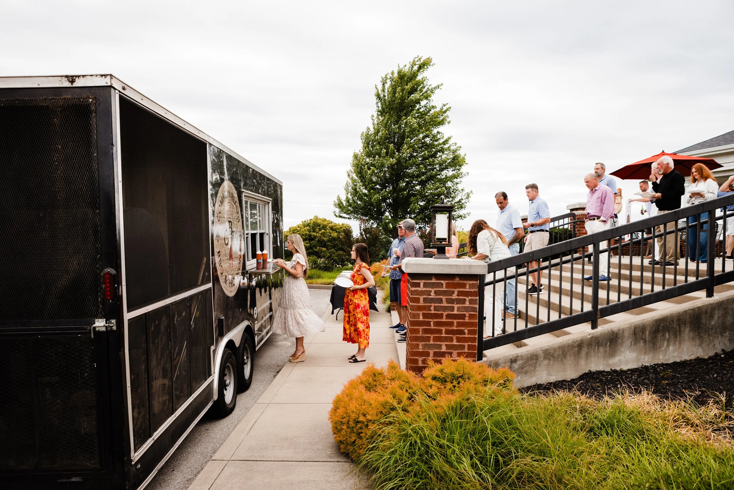 People standing in line outside a food truck on a cloudy day. The food truck is black with a window serving customers. Some people are holding plates or drinks, waiting on steps leading up to a building. There are trees, bushes, and a brick railing in the background.