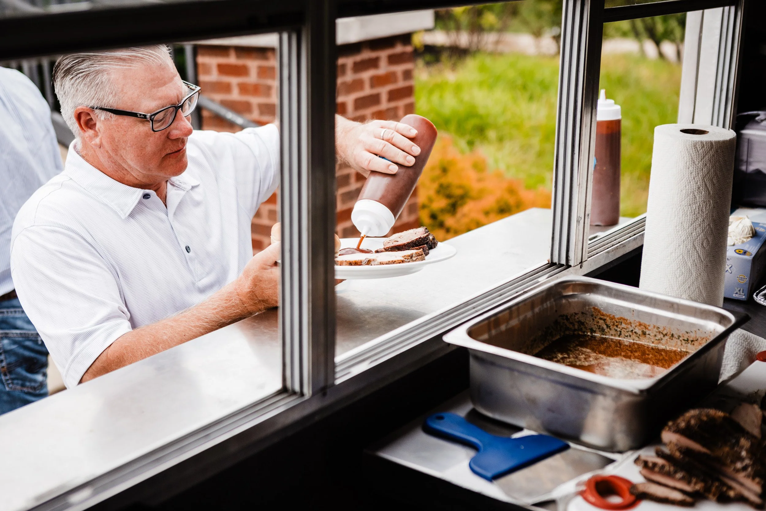 A man with gray hair and glasses is pouring sauce onto slices of meat or brisket on a plate. The scene takes place at a food stand or truck with a brick wall background. Various food prep items are visible on a counter.