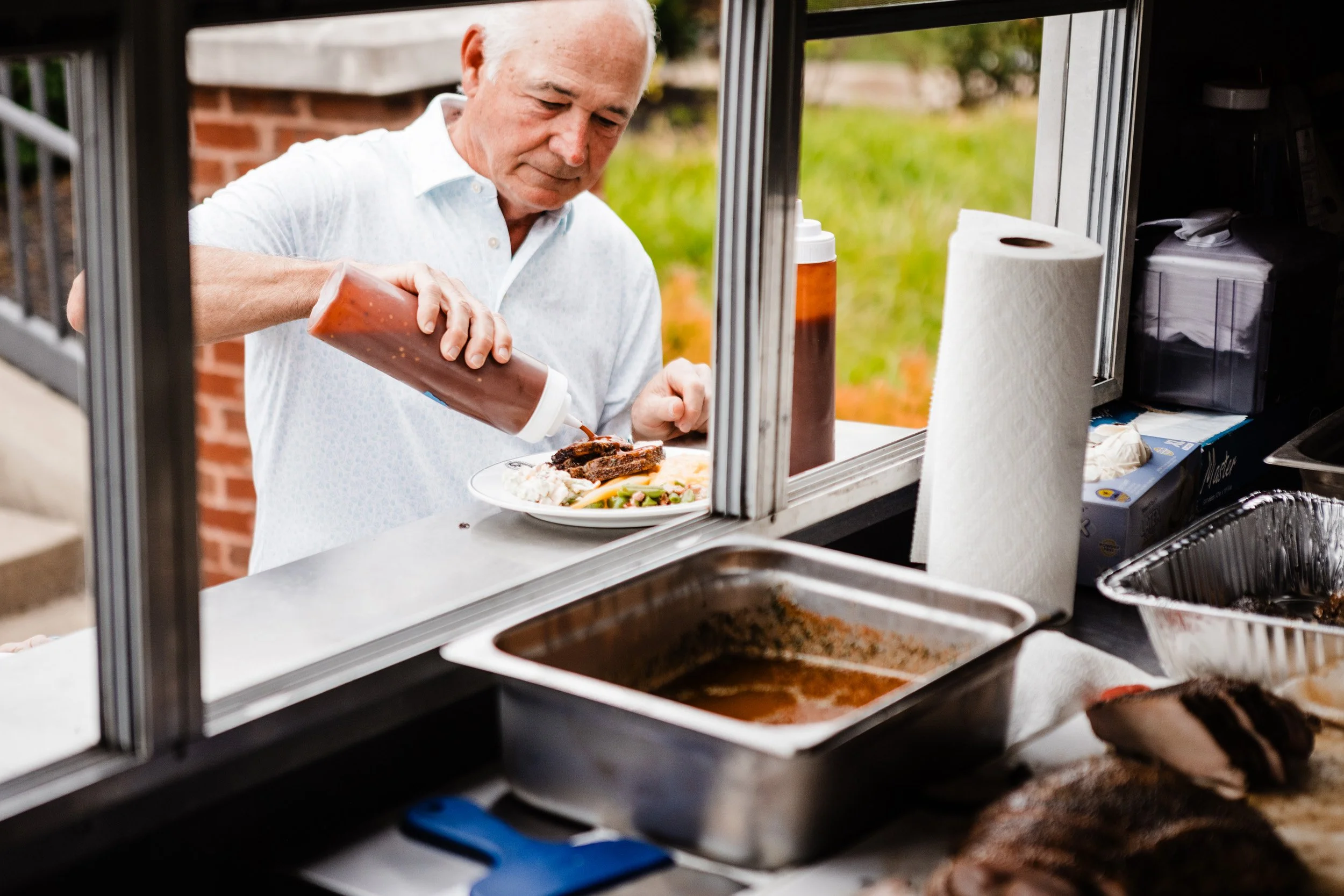 An elderly man is preparing food at a food truck or outdoor kitchen, pouring sauce on a plate of food that includes meat and vegetables. The setting is outdoors with greenery and a brick wall in the background.