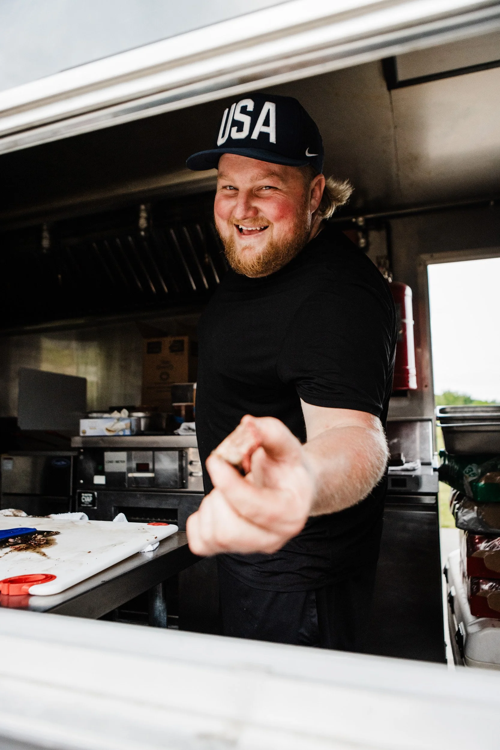 Smiling chef in a USA hat offering a piece of BBQ meat from the window of a food truck.