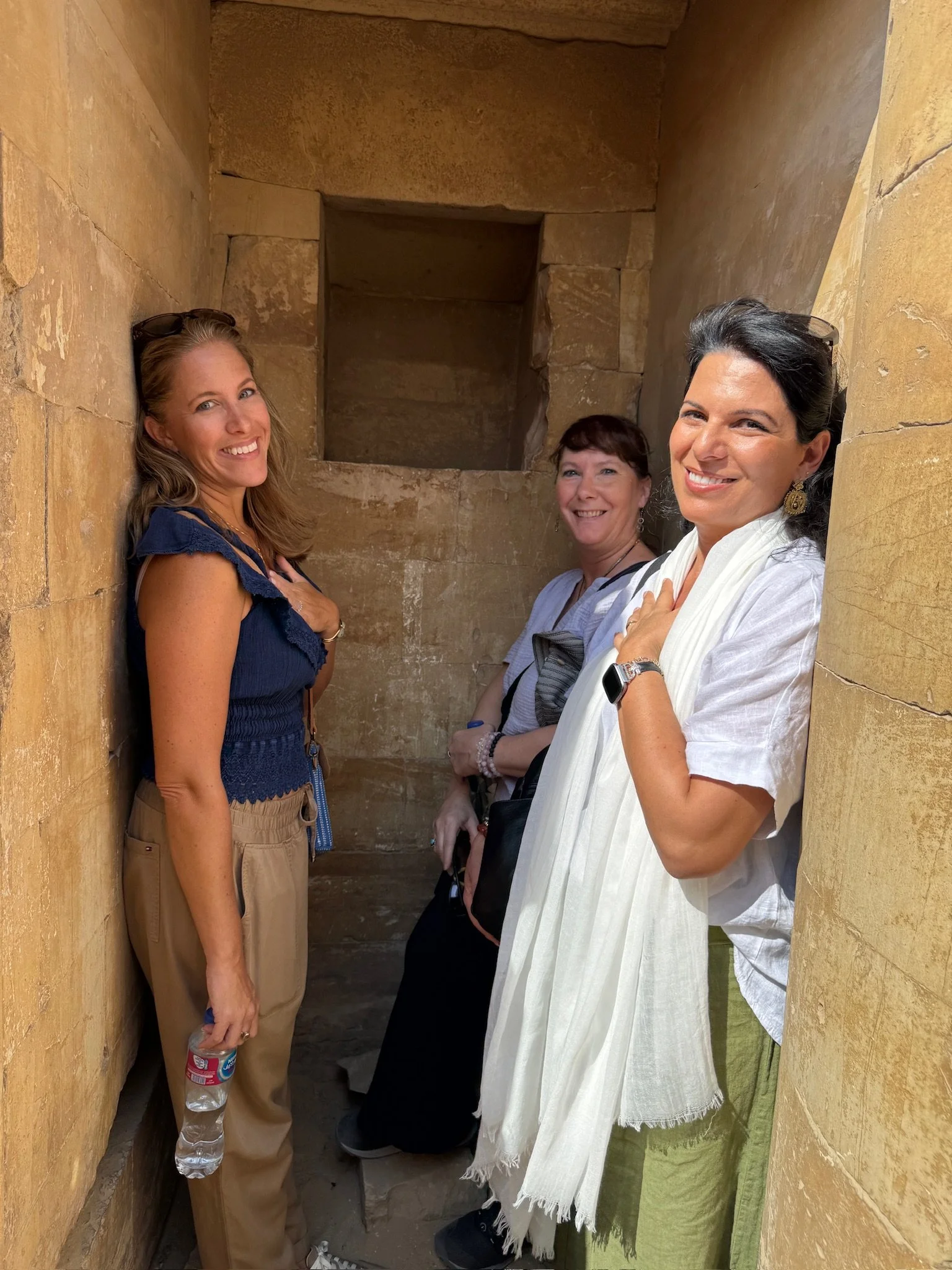 Three women standing in a narrow stone corridor with rough stone walls, smiling at the camera.