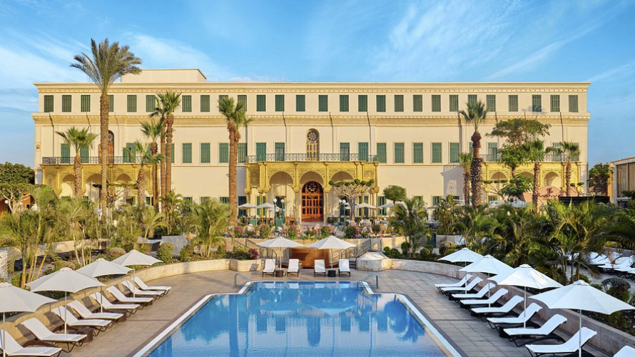 Luxury hotel with a swimming pool, palm trees, and a grand beige building in the background under a blue sky
