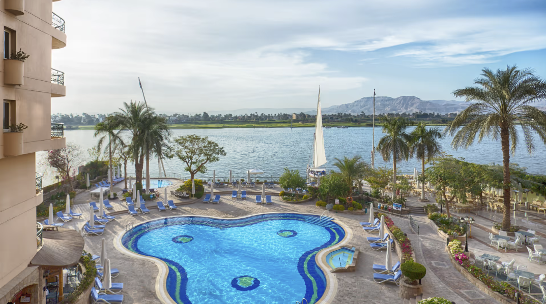 Scenic view of a hotel pool area with lounge chairs and umbrellas, overlooking a lake with sailboats, palm trees, and mountains in the background.