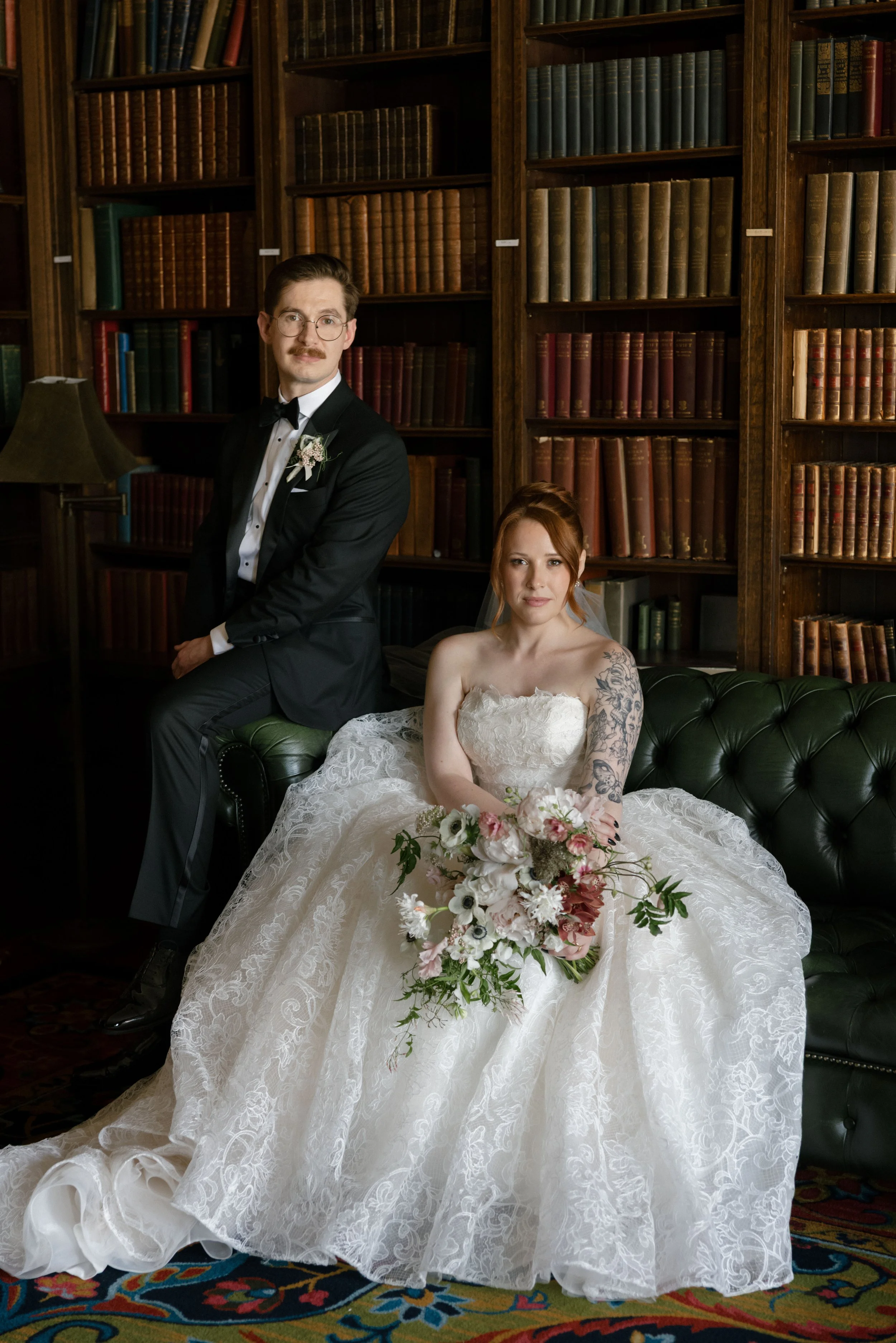 A bride in a white wedding gown sitting on a green leather couch holding a bouquet, with a groom in a tuxedo standing beside her in a wood-paneled library with bookshelves filled with books.