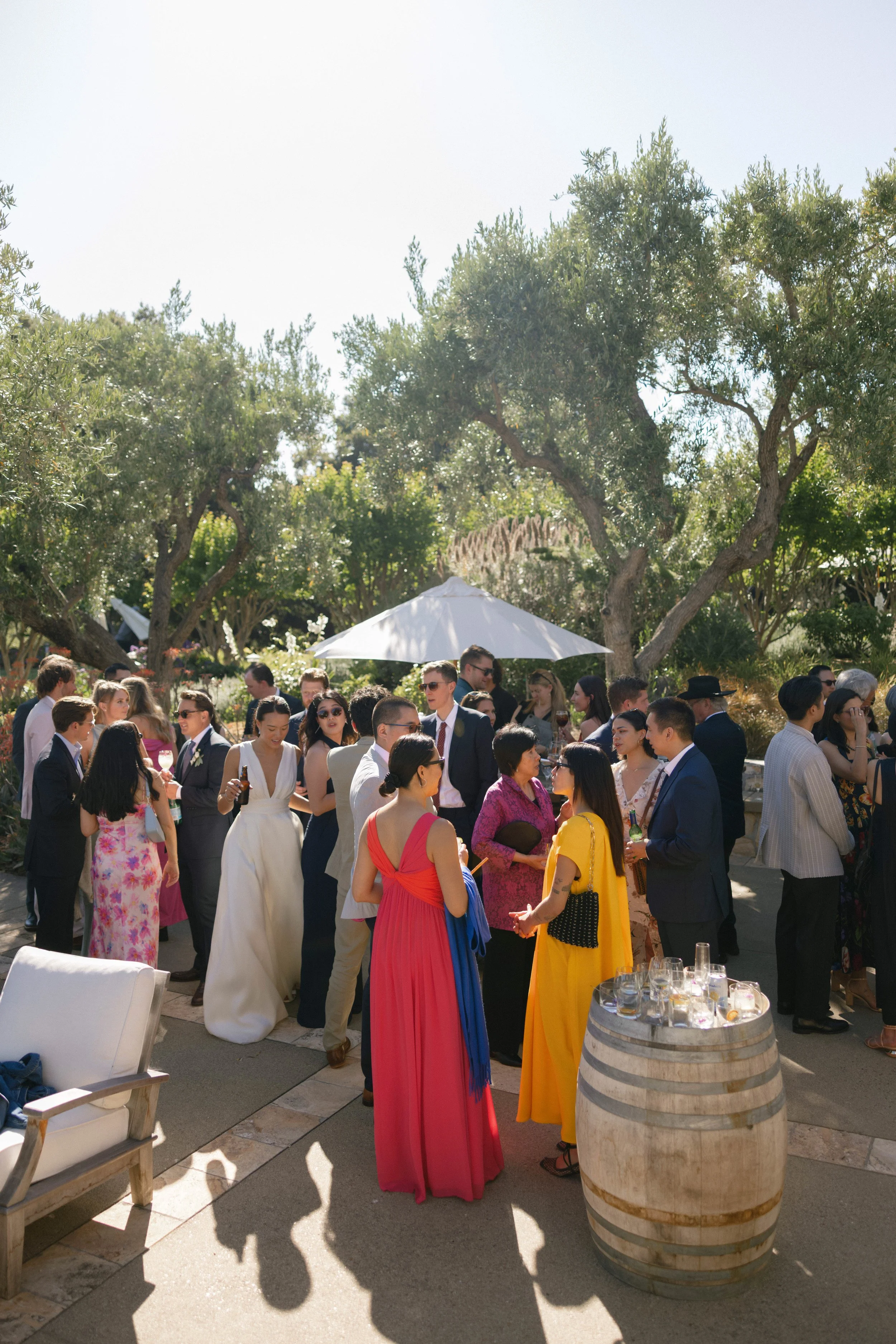 Group of people at an outdoor event, socializing under trees with a white umbrella, on a sunny day.