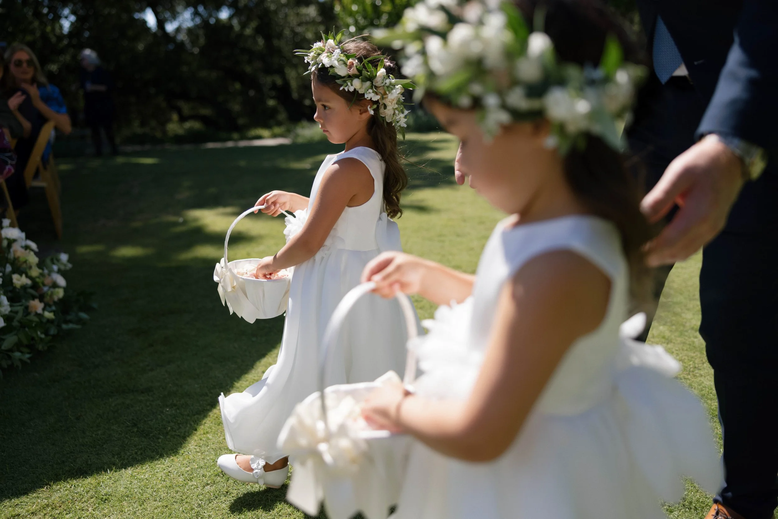Two young girls dressed in white dresses with flower crowns on their heads, holding baskets, participating in a wedding or similar outdoor ceremony.