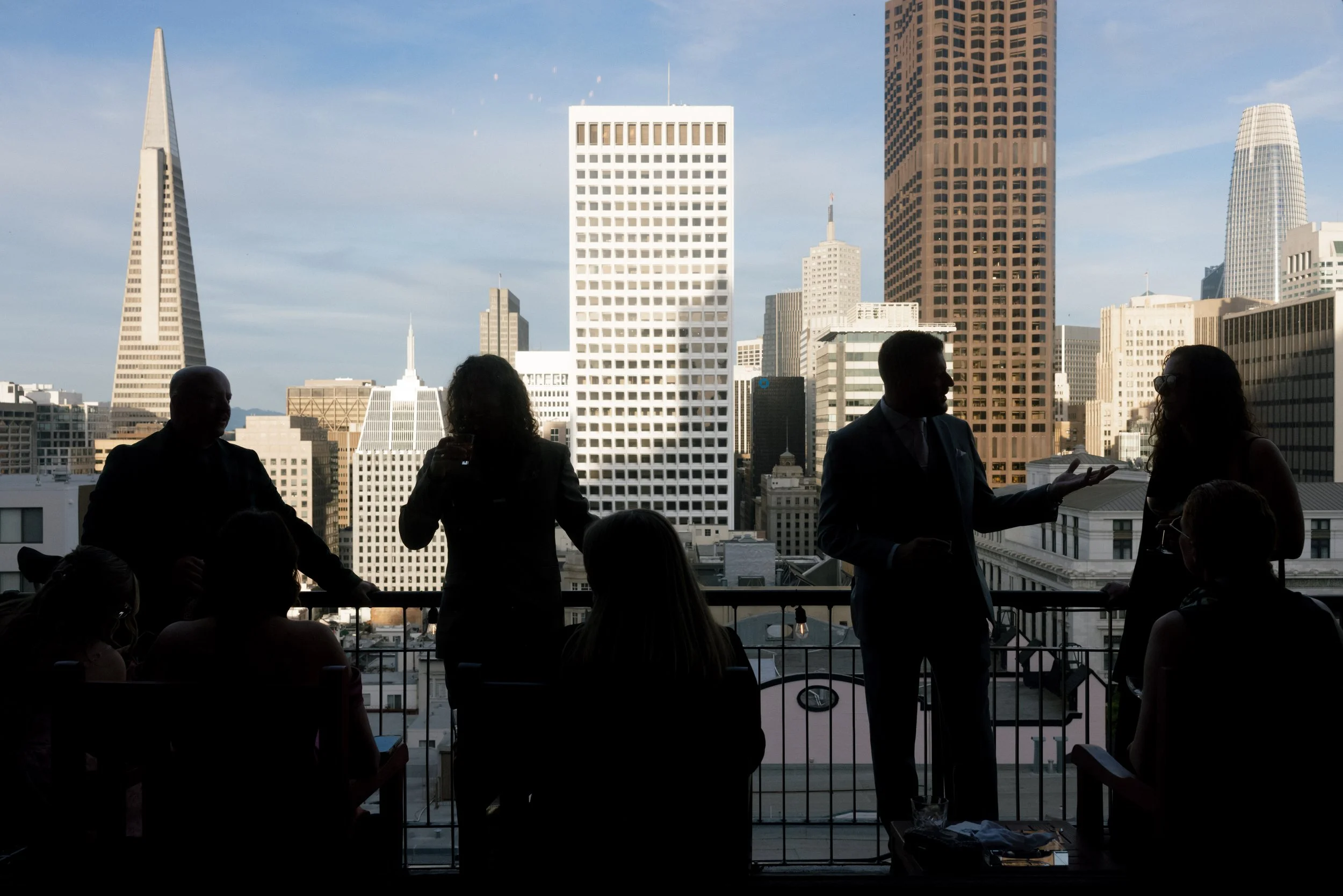 Silhouetted group of people on a balcony with a city skyline featuring tall buildings and skyscrapers in the background during daytime.