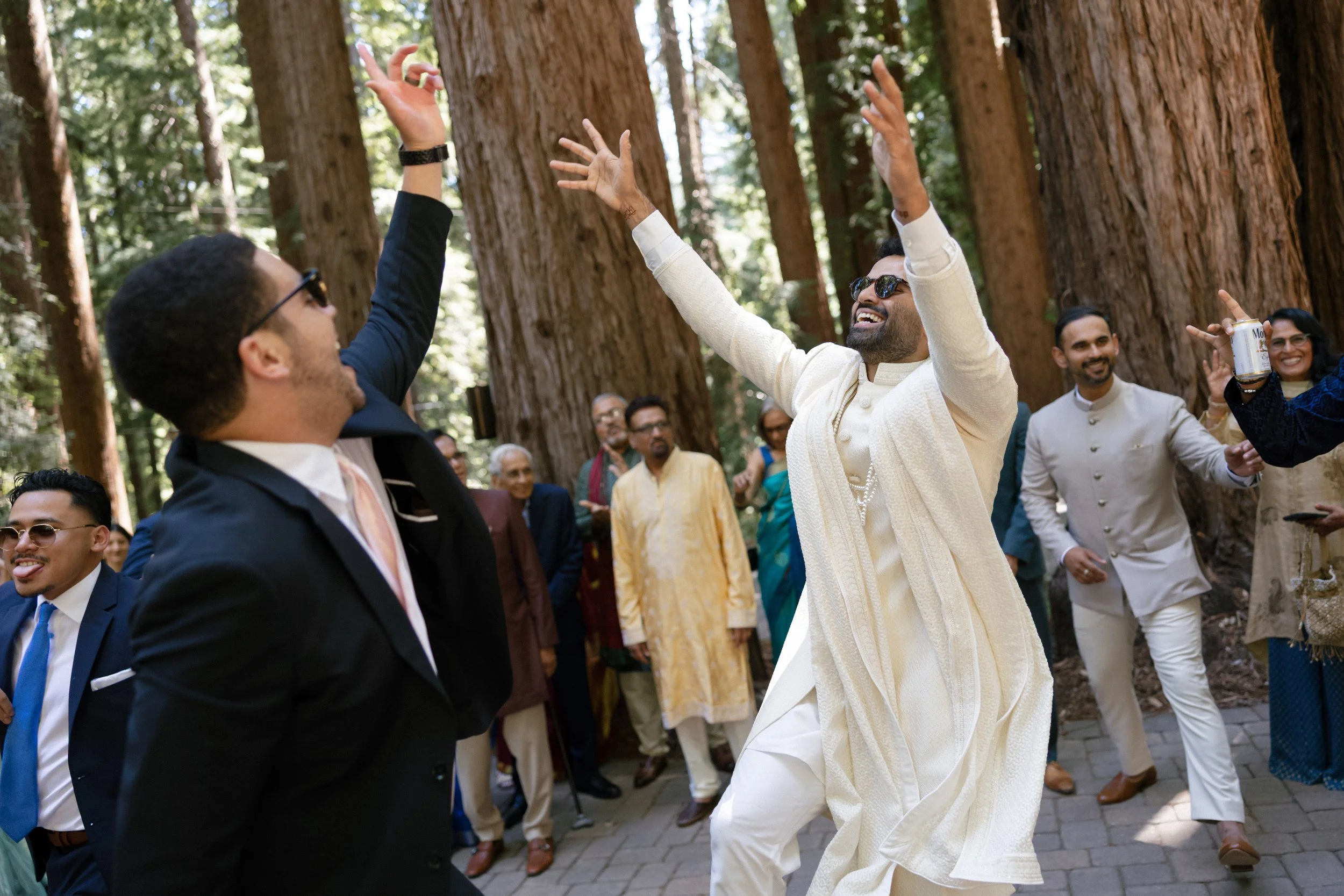 A group of people dancing and celebrating outdoors among tall trees during a festive event, with some dressed in traditional Indian attire.