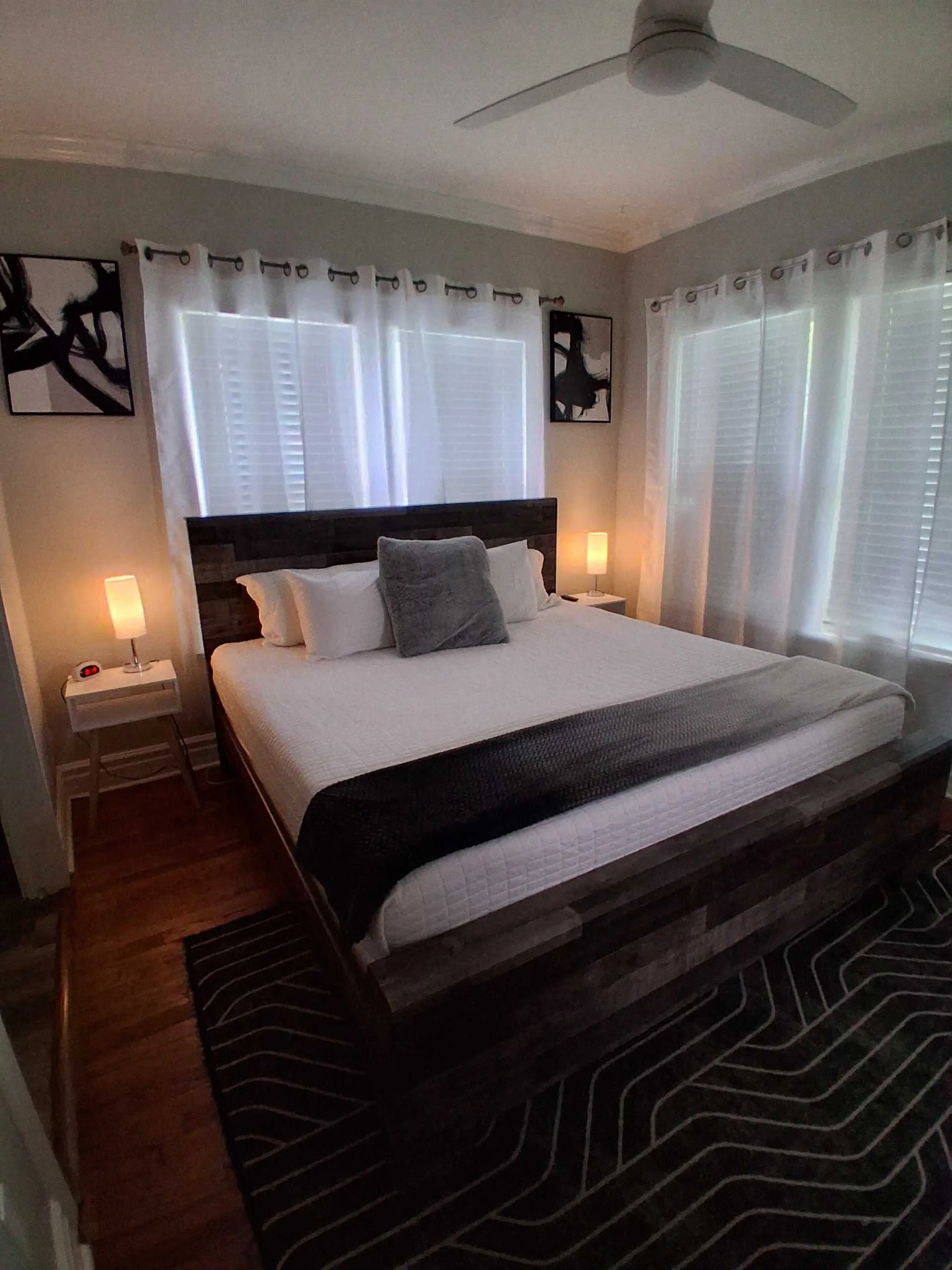 A bedroom featuring a bed with white bedding and a dark headboard, a gray pillow, two small white bedside tables with lamps, large windows with white blinds and curtains, two abstract black and white art pieces on the wall, a ceiling fan, and a patterned dark rug on a wooden floor.