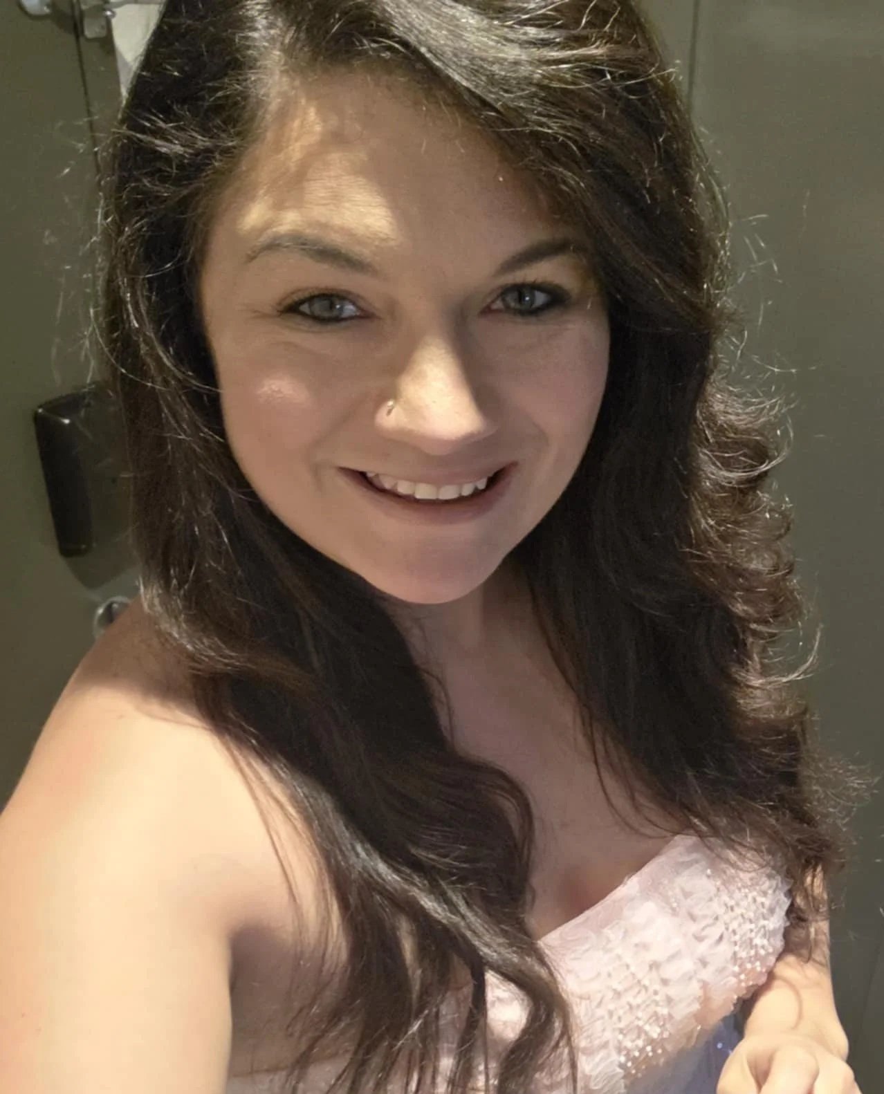 Close-up of a woman with long, wavy brown hair, blue eyes, and a nose piercing, smiling at the camera, wearing a light-colored strapless dress.
