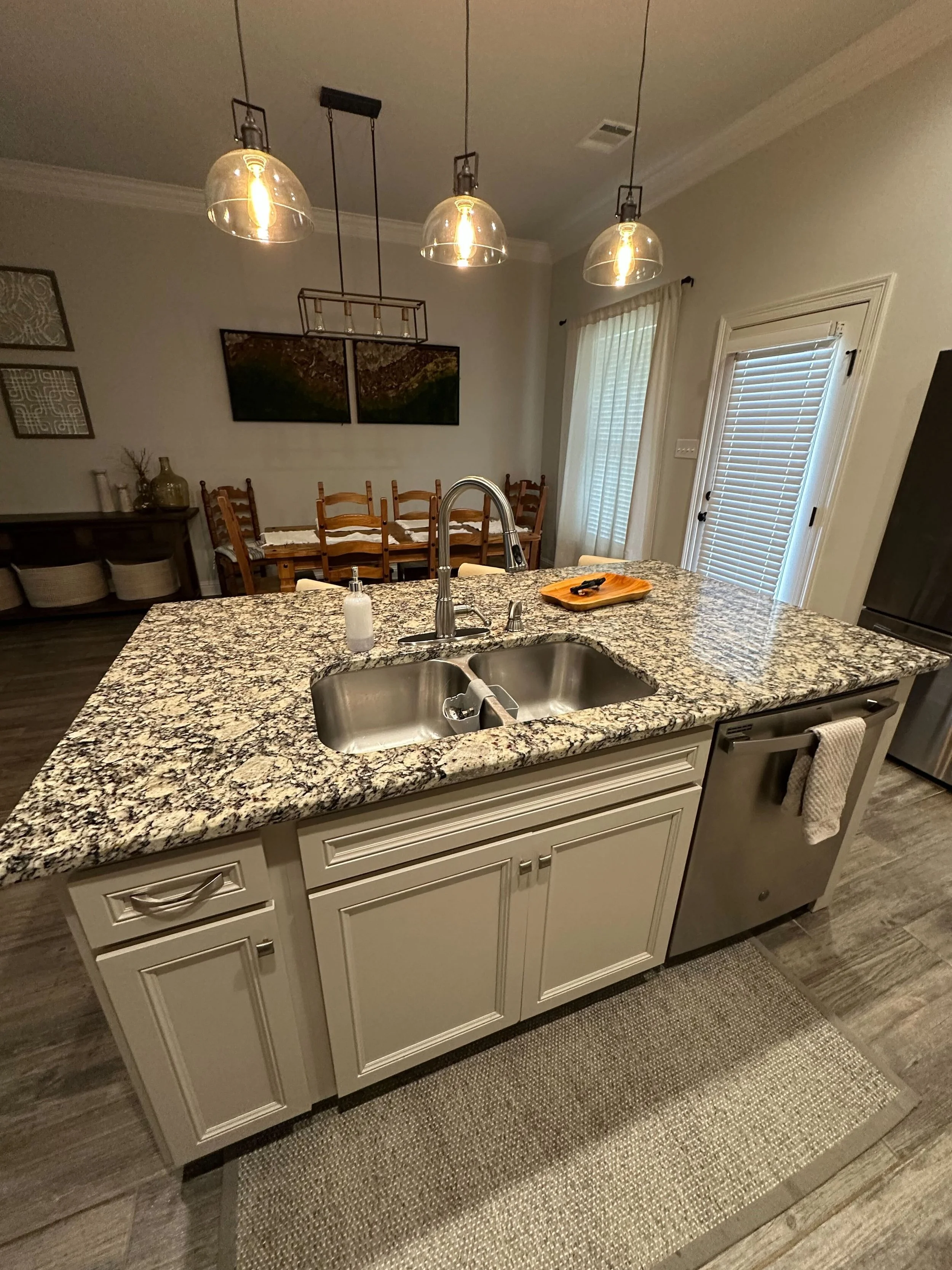 Kitchen island with granite countertop, stainless steel sink, and a soap dispenser, with a beige cabinet underneath, and a beige rug on a wooden floor.