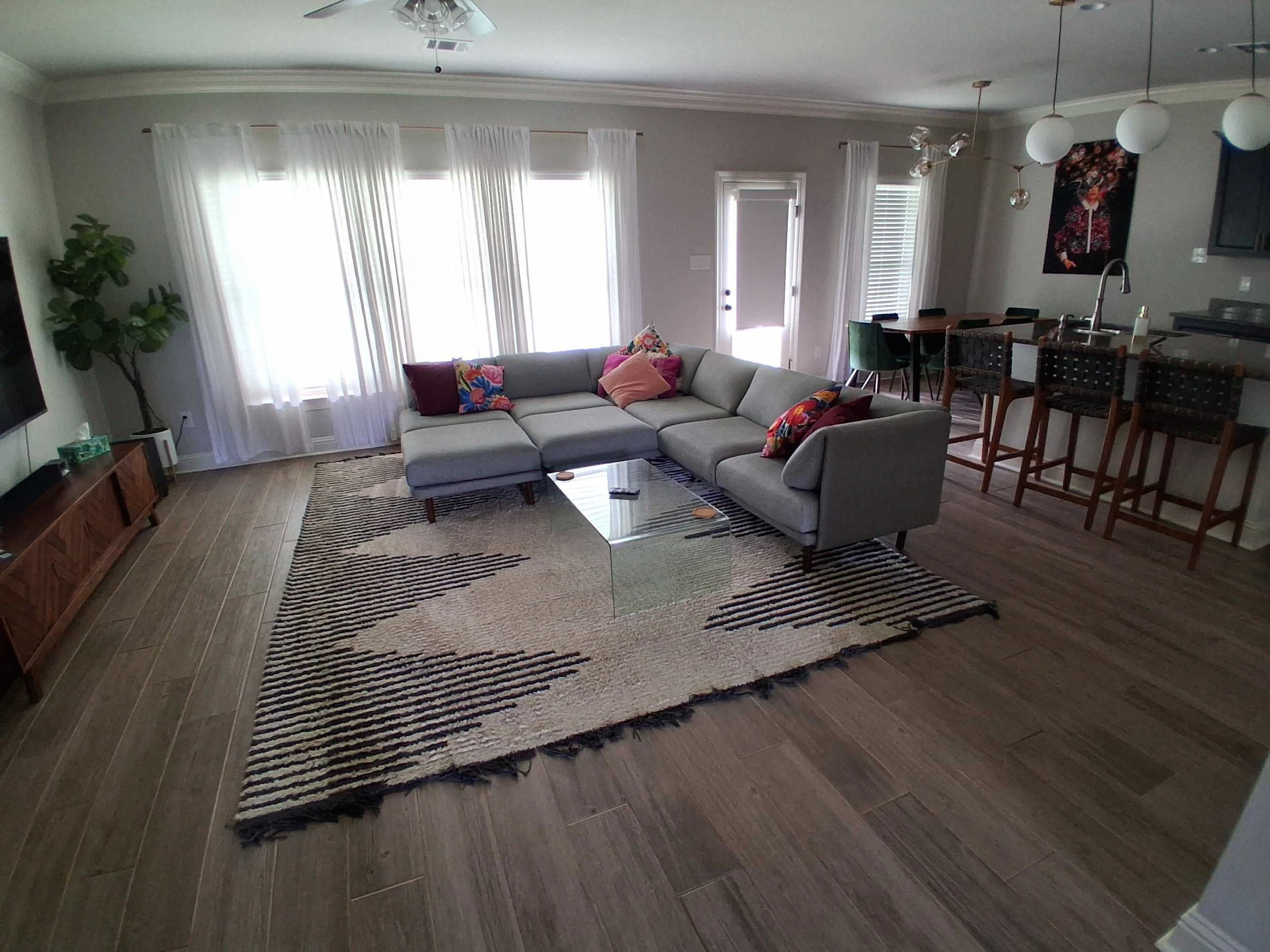 Living room with gray sectional sofa, colorful throw pillows, wooden coffee table, patterned rug, large windows with white curtains, potted plant, dining area with table and chairs, kitchen island with barstools, modern lighting fixtures.