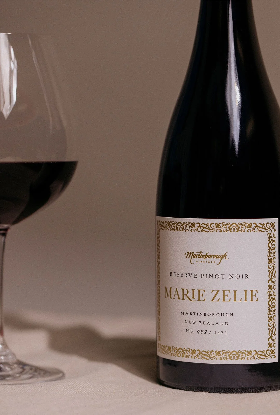 A bottle of Marie Zelie Reserve Pinot Noir wine from Martinborough, New Zealand, next to a glass of red wine on a light-colored table.