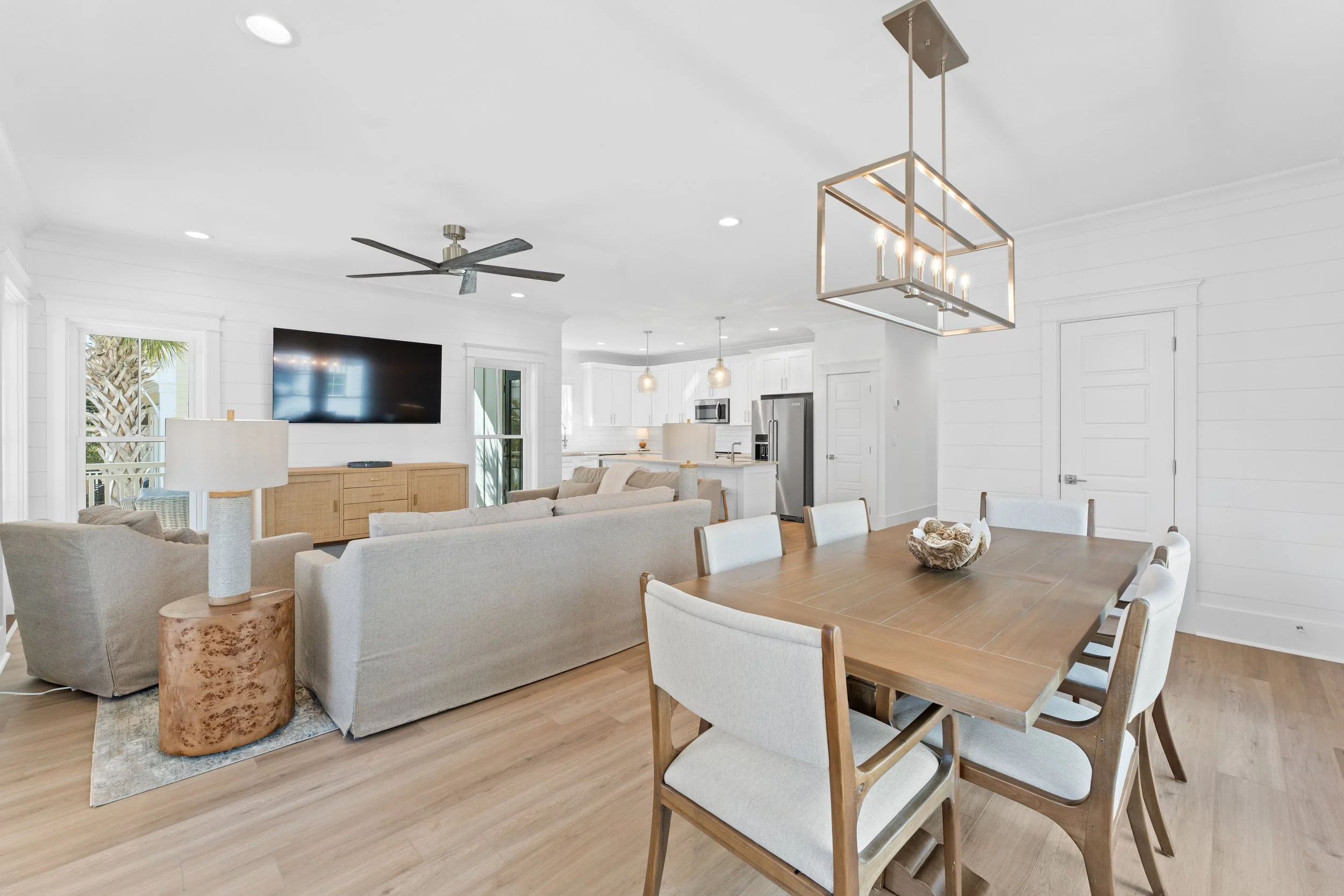 Open-concept living and dining area with modern decor, white walls, hardwood floors, sectional sofa, dining table with eight chairs, kitchen with white cabinets and stainless steel appliances, ceiling fan, pendant lights, and large windows letting in natural light.