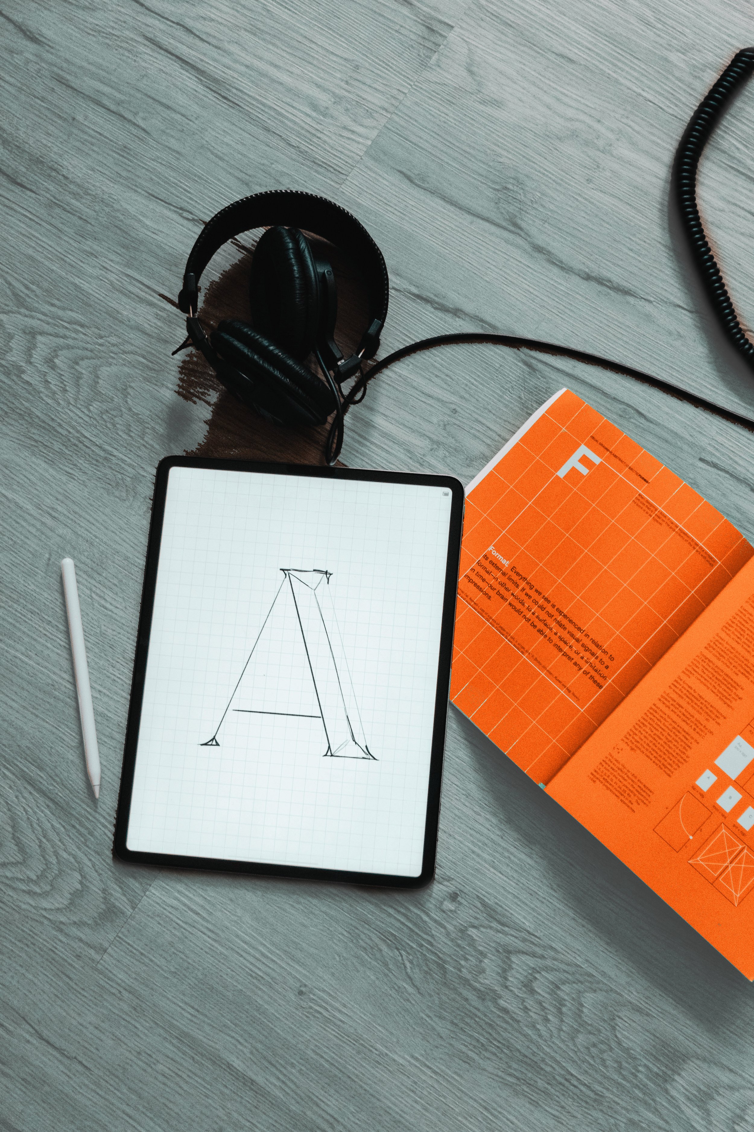 A digital tablet displaying a hand-drawn sketch of the letter 'A', surrounded by a stylus, headphones, and an orange technical magazine on a wooden floor.