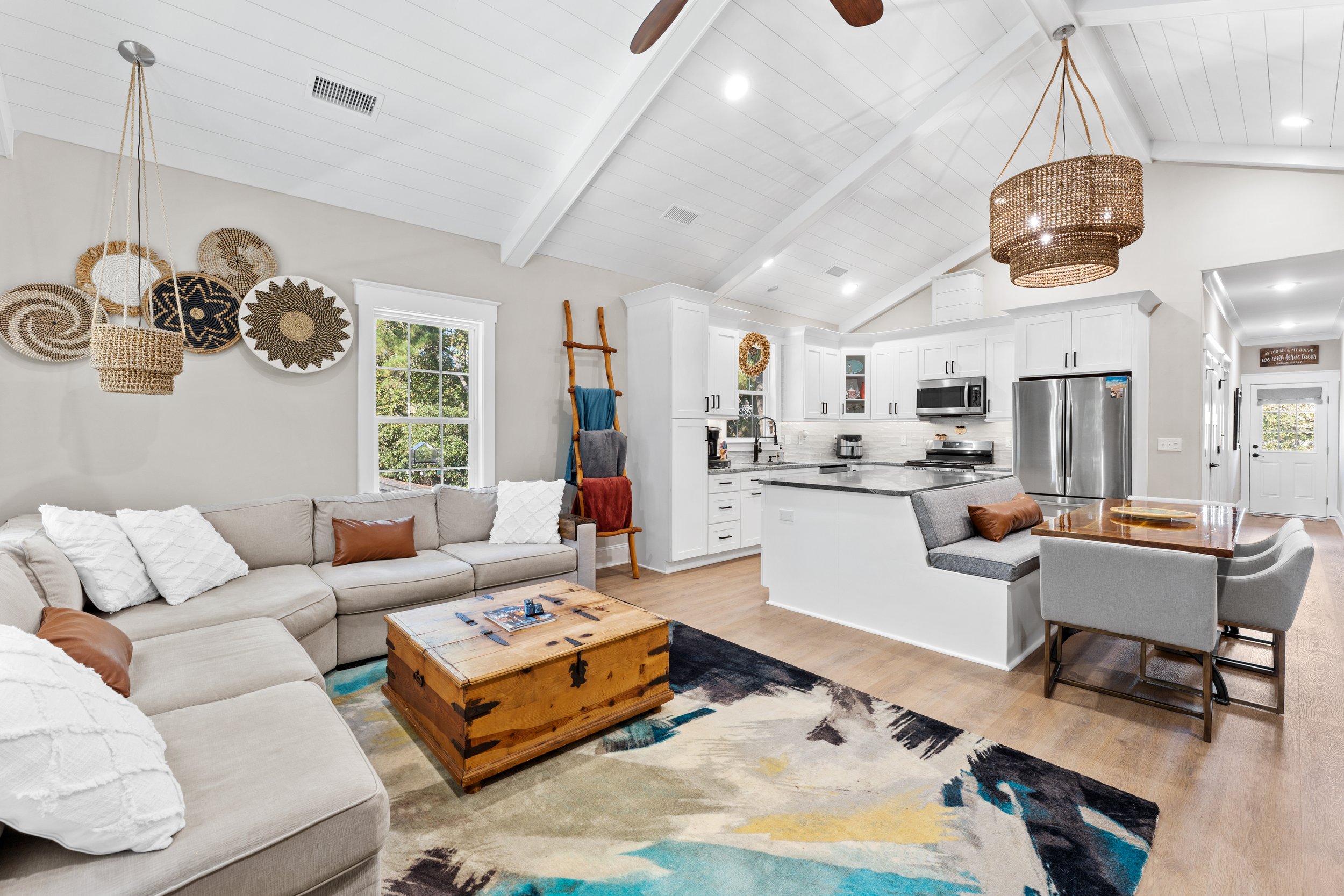 Open-concept living room and kitchen with white walls, vaulted ceiling with wooden beams, and large windows. The living area has a beige sectional sofa with white and brown pillows, a wooden coffee table, and wall decor framed baskets. The kitchen features white cabinets, stainless steel refrigerator, microwave, and a breakfast bar with gray upholstered seating. Light hardwood floors and woven pendant lights hang from the ceiling.