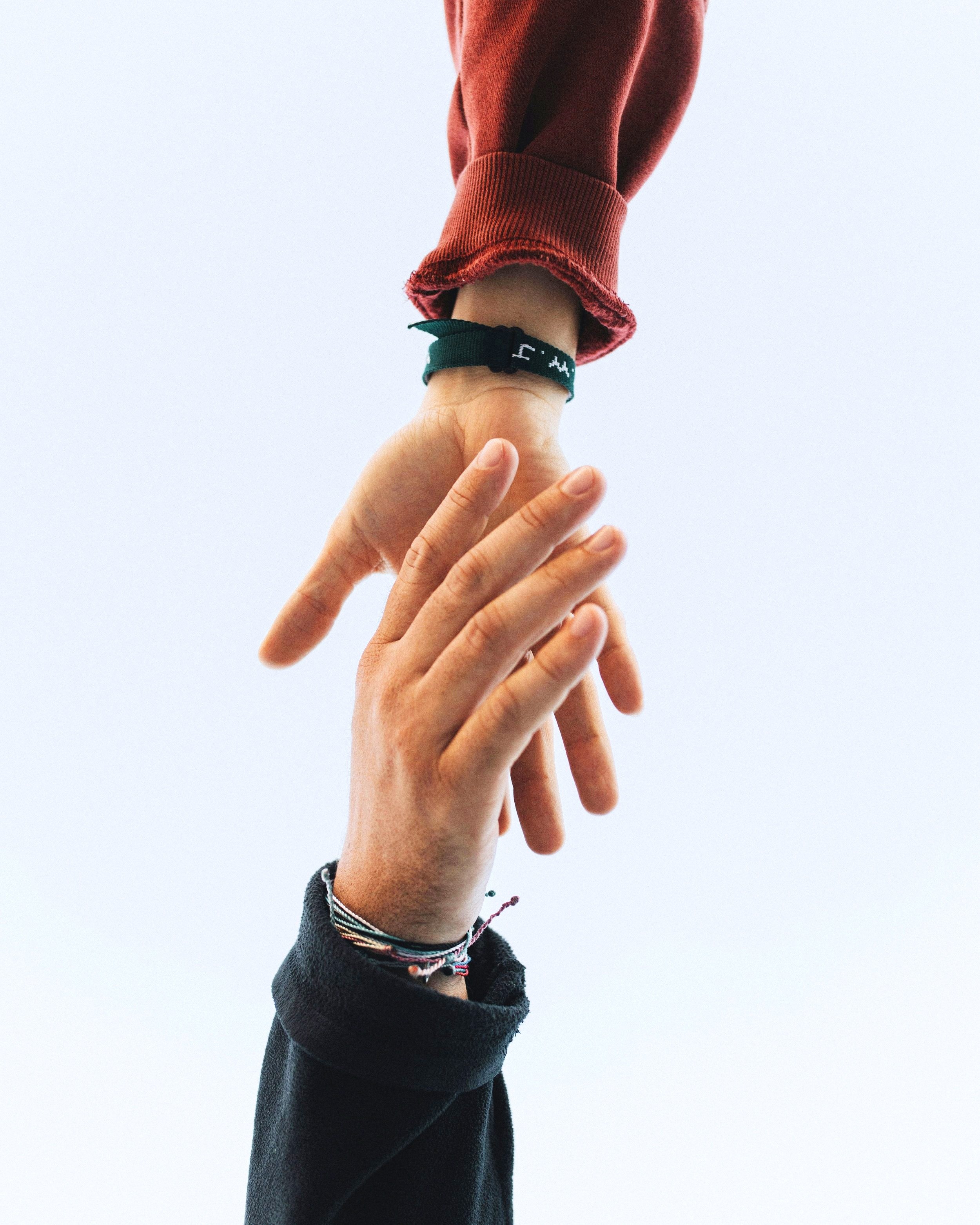 Two hands reaching towards each other against a plain background, one hand wearing a black long-sleeve shirt with bracelets, the other hand wearing a red long-sleeve shirt with a wristband.