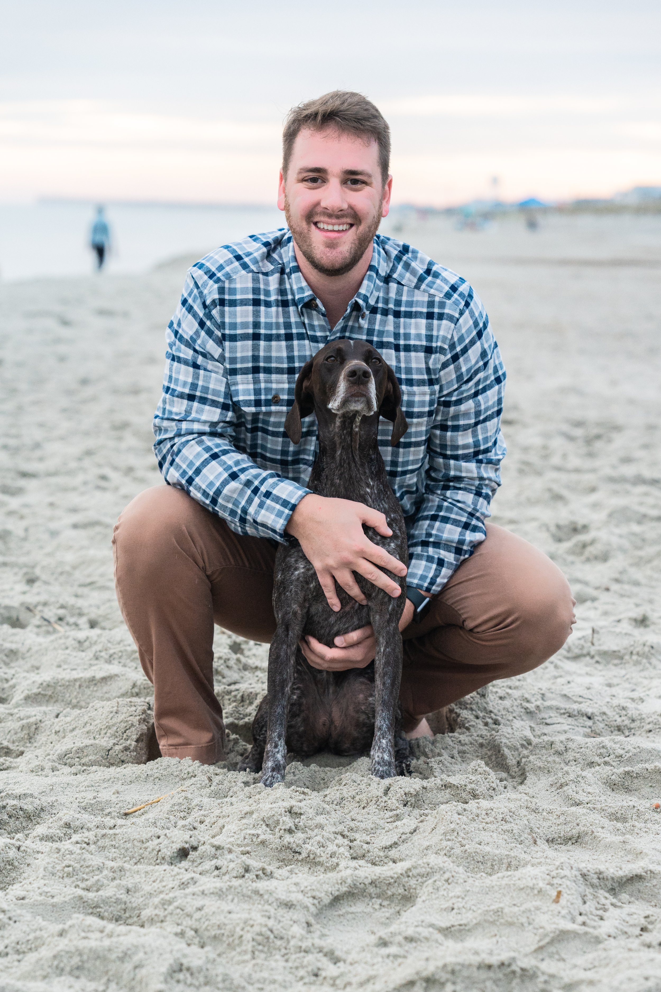 A man in a plaid shirt squatting on a sandy beach, holding a black and brown dog, with the ocean and a person walking in the background during sunset.
