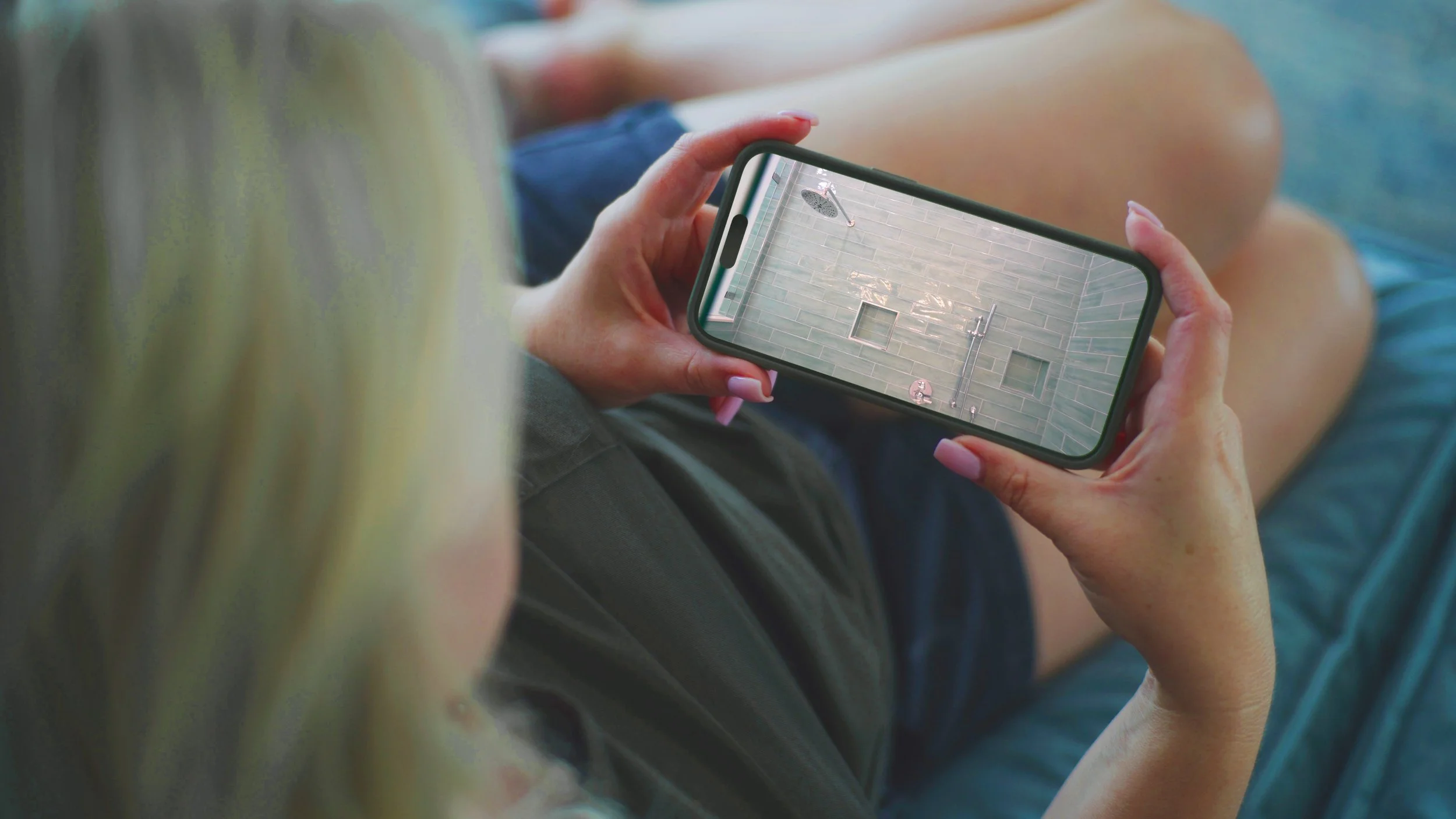 A person with long blonde hair sitting and looking at their phone, which shows a video game scene with a tiled wall, showerhead, and faucet.