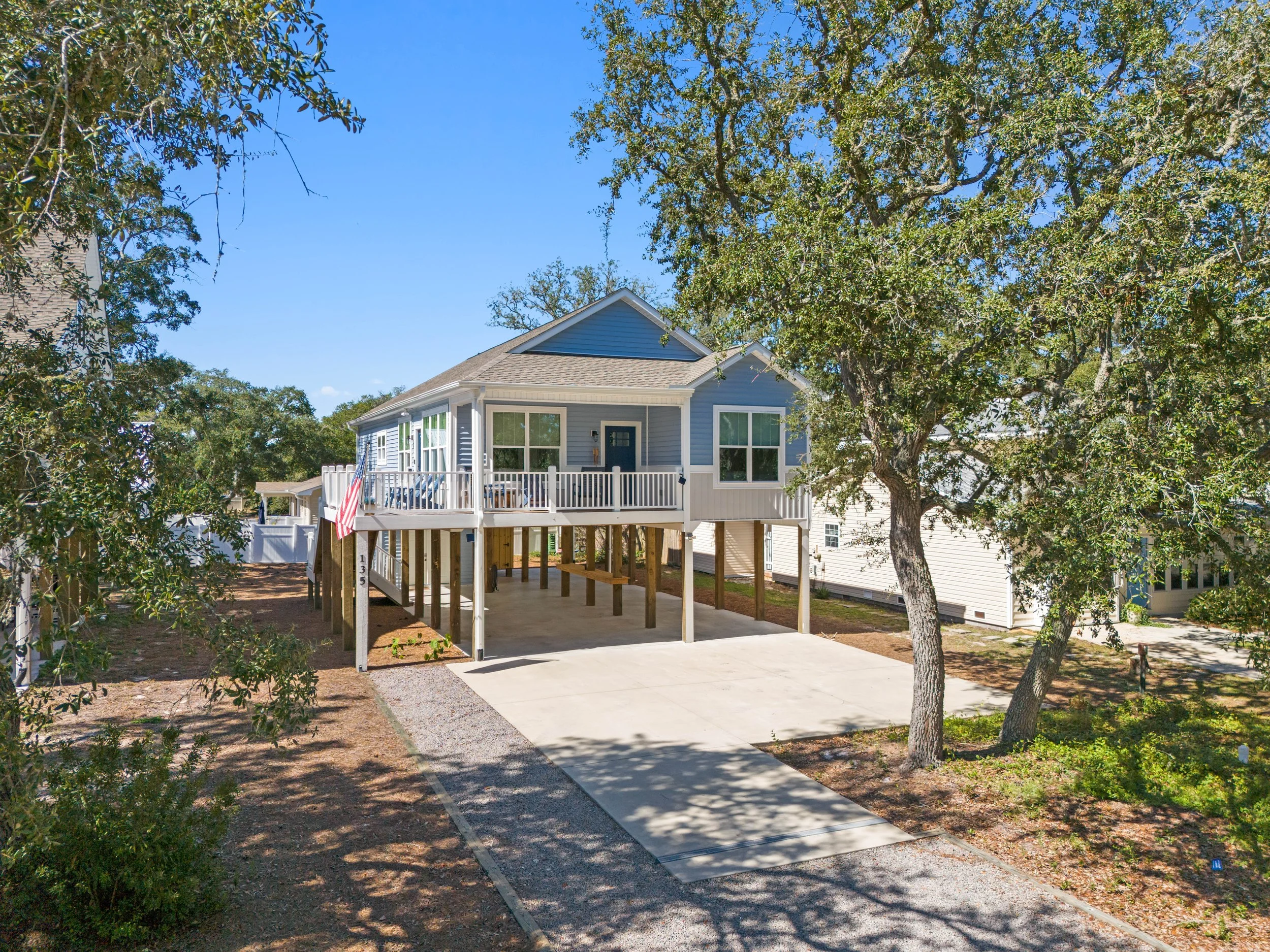 A two-story blue house with a large porch and white railings, elevated on stilts, with a concrete driveway leading up to it, surrounded by trees and a clear blue sky.