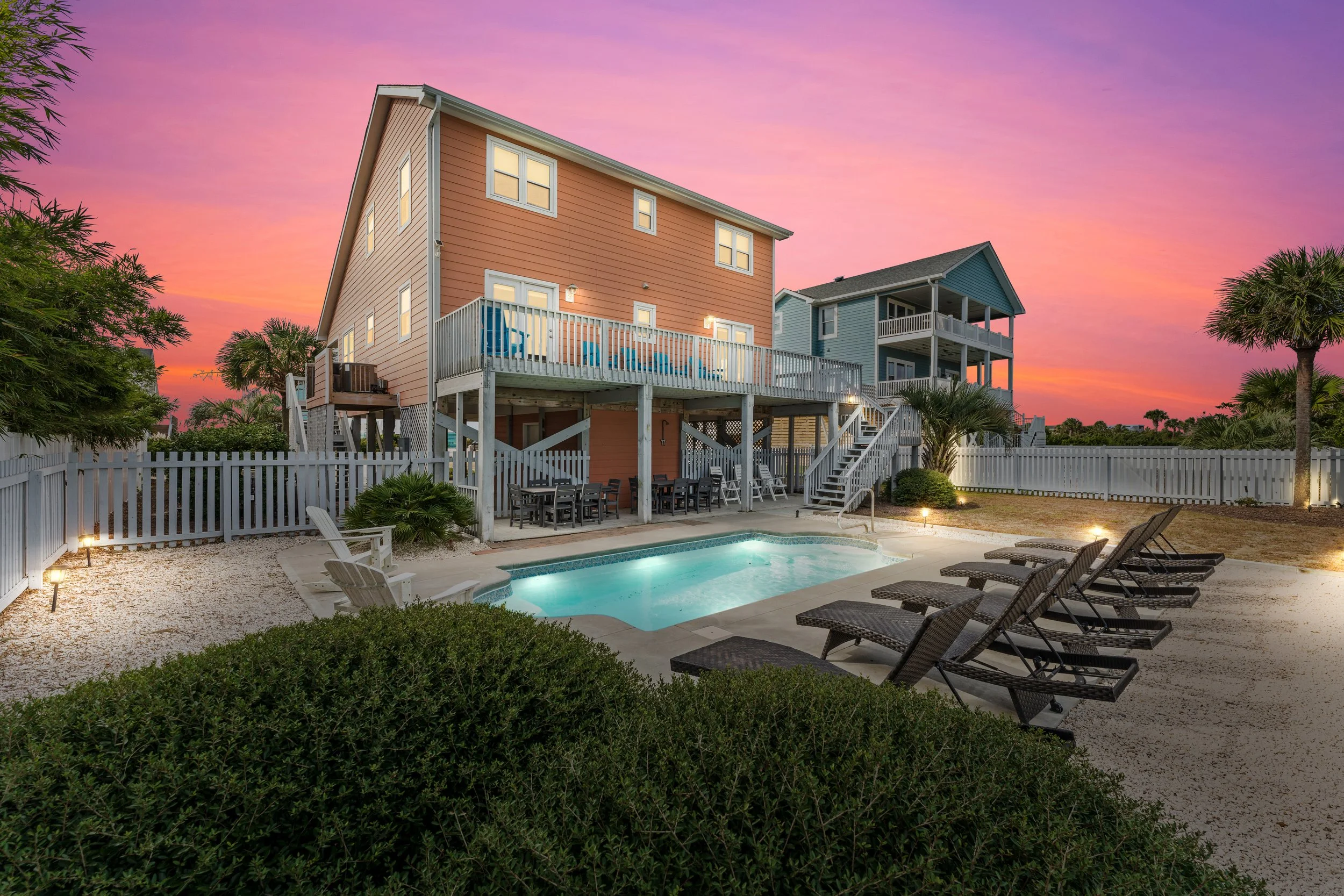 A backyard pool area with pool chairs, a deck, and two houses during sunset with a pink and purple sky.