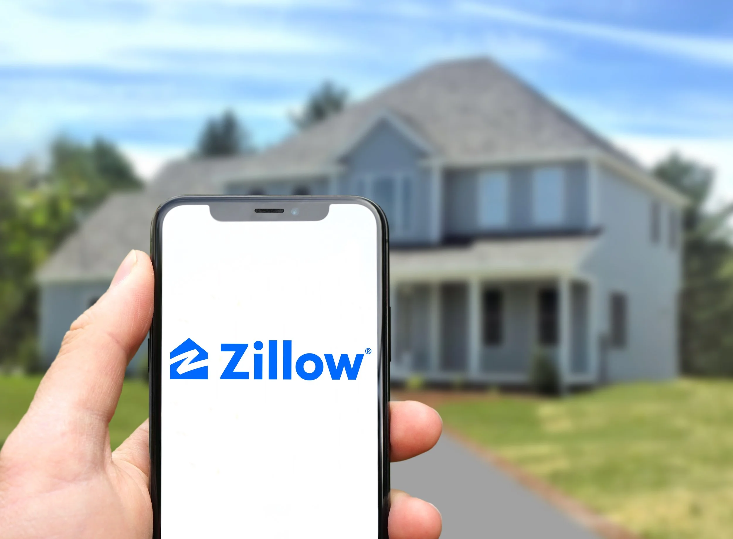 Hand holding a smartphone with the Zillow app logo in front of a house.