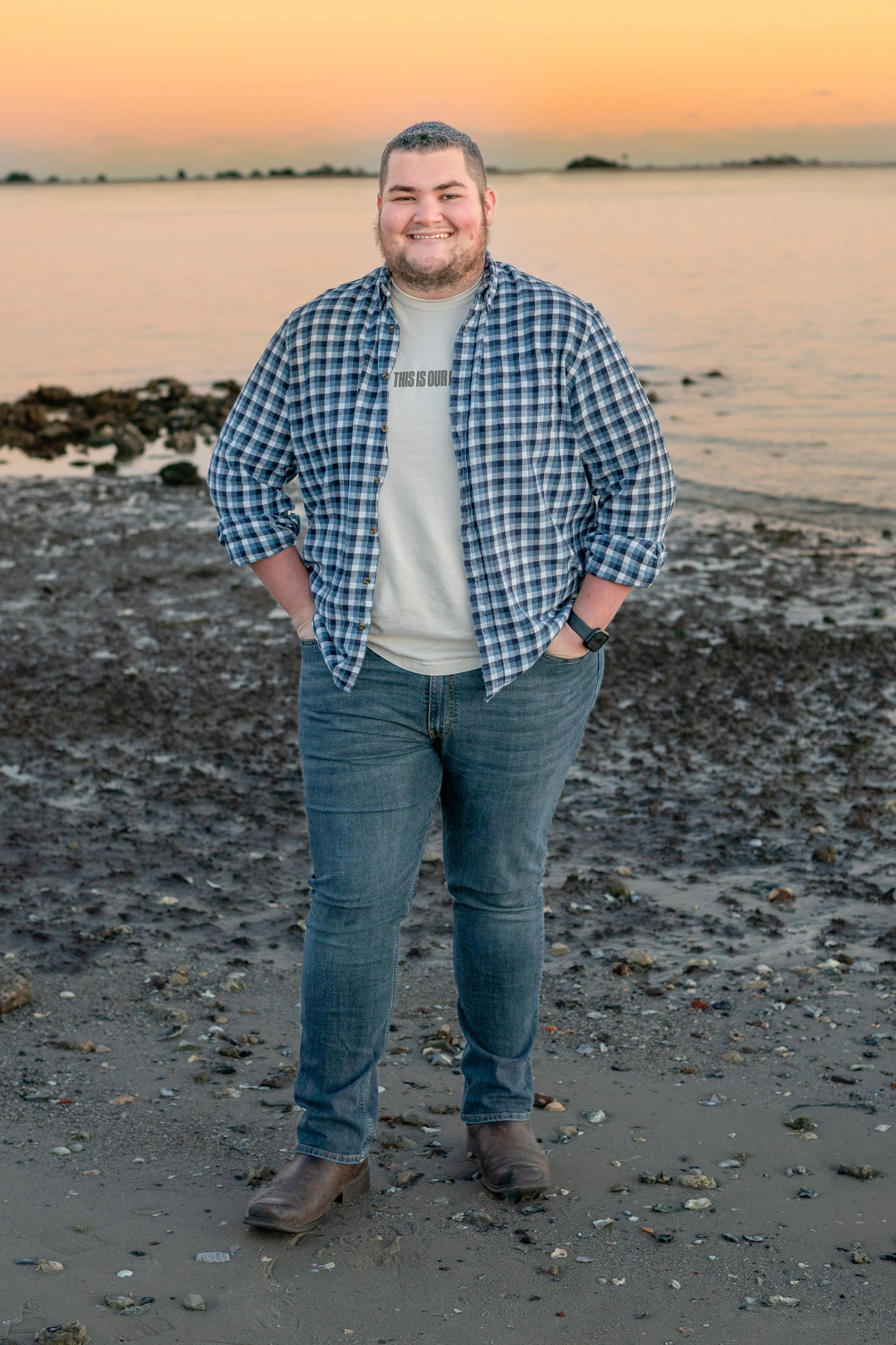 A man stands on a rocky beach at sunset, wearing a plaid shirt, jeans, and boots, smiling at the camera.