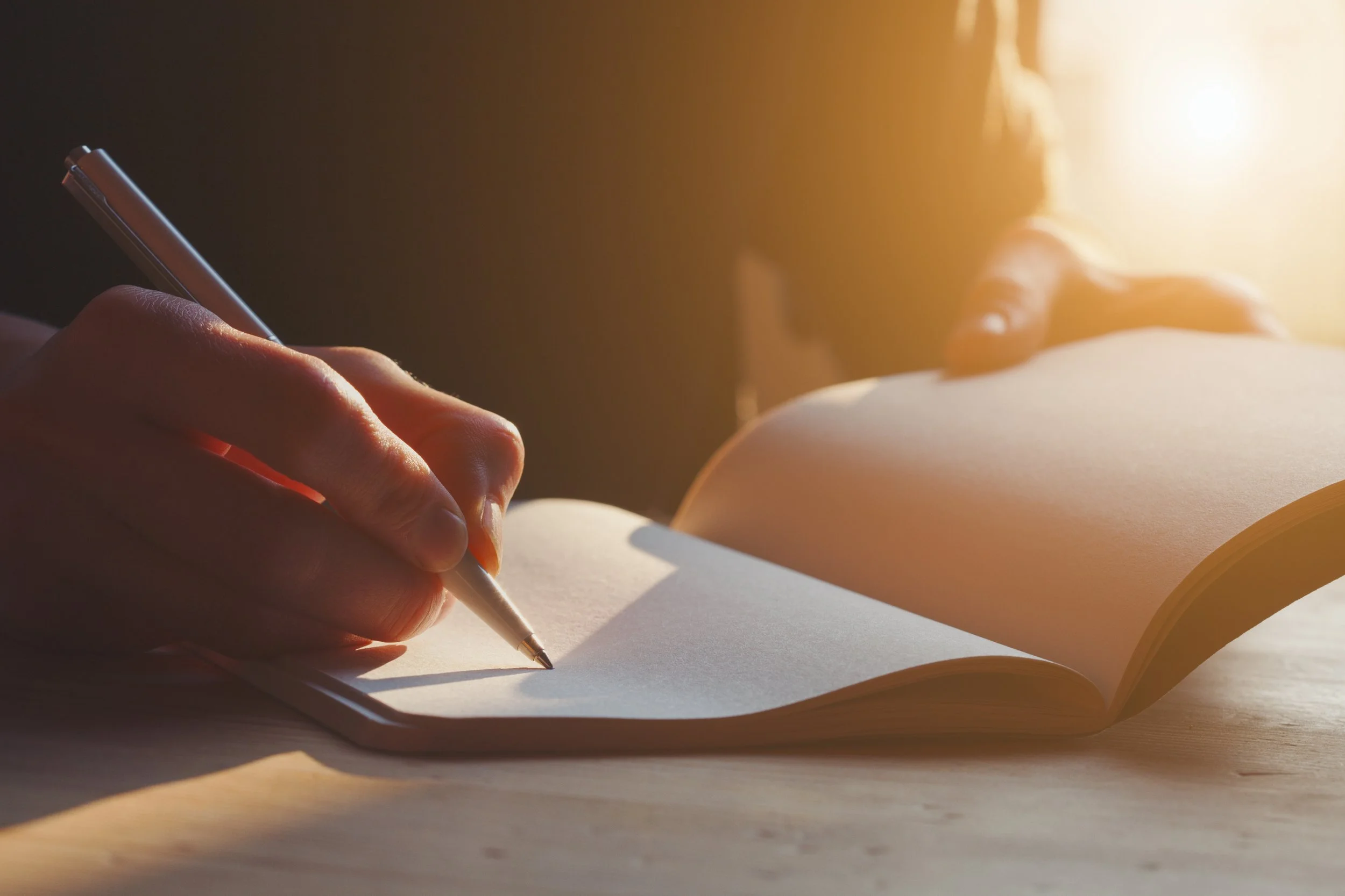 Close-up of a person writing in a notebook with a ballpoint pen, sunlight streaming in from the right.