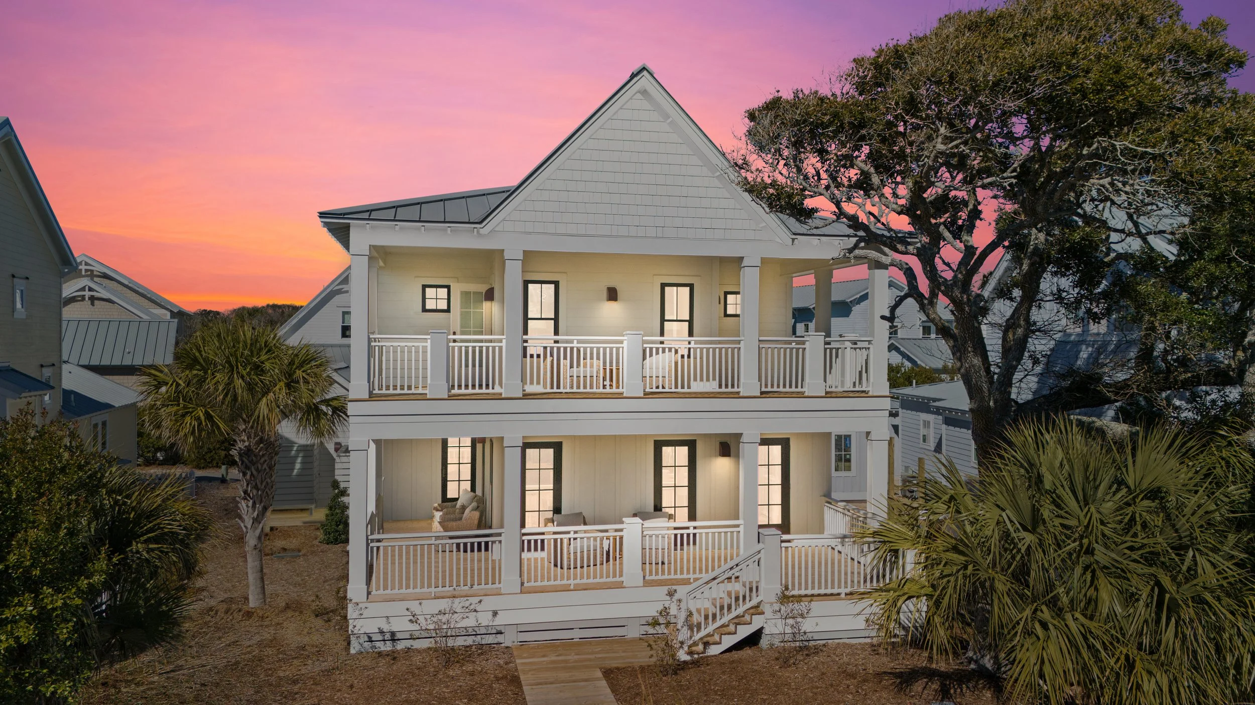 A two-story house with front and back porches, white railings, and exterior wall lights, surrounded by trees and neighboring houses, during sunset with pinkish-purple sky.